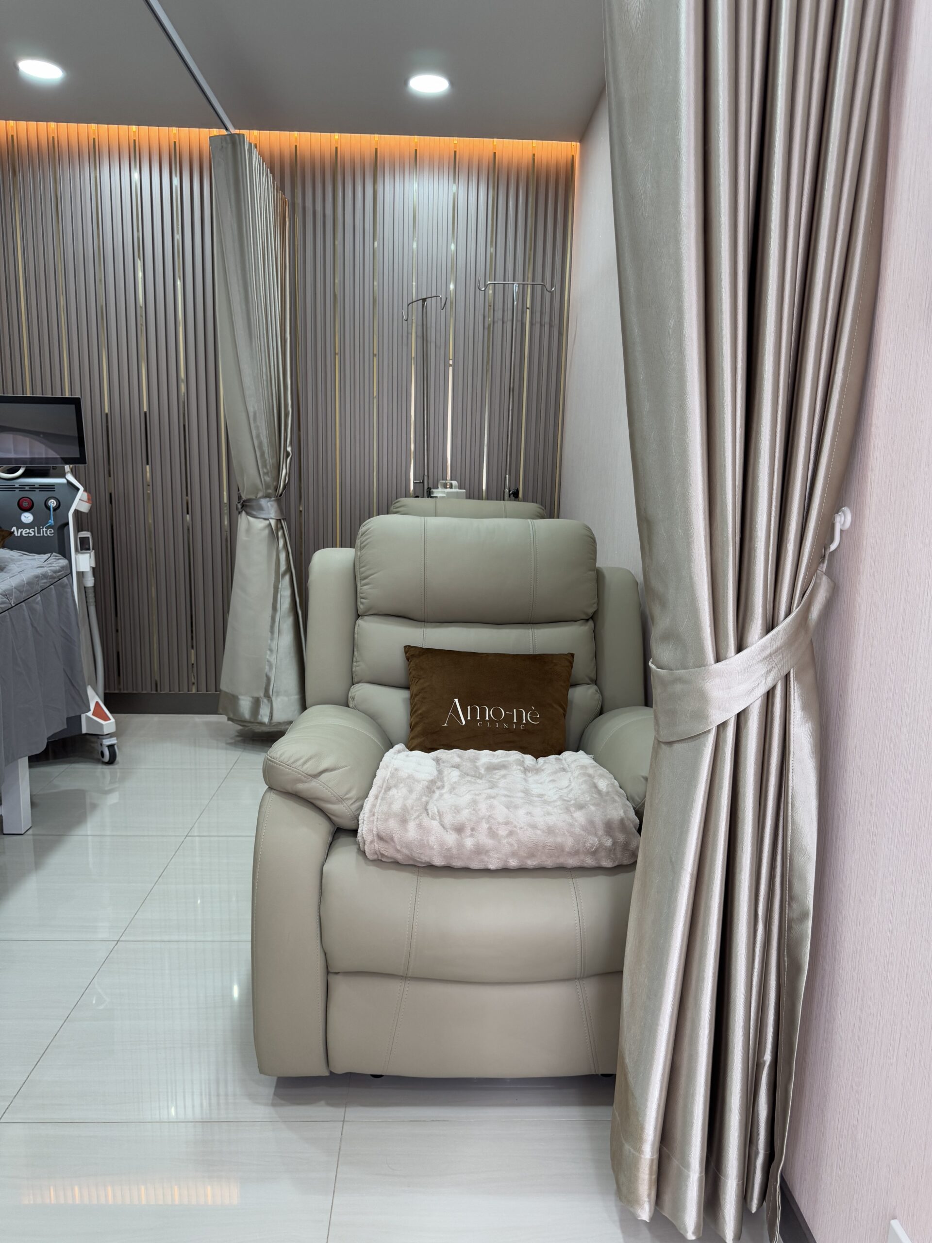 เก้าอี้ปรับเอนหนังสีครีมพร้อมเสาแขวนน้ำเกลือในห้องทรีทเมนต์คลินิก Amo-nè ตกแต่งด้วยผ้าม่านสีเงินและไฟ LED