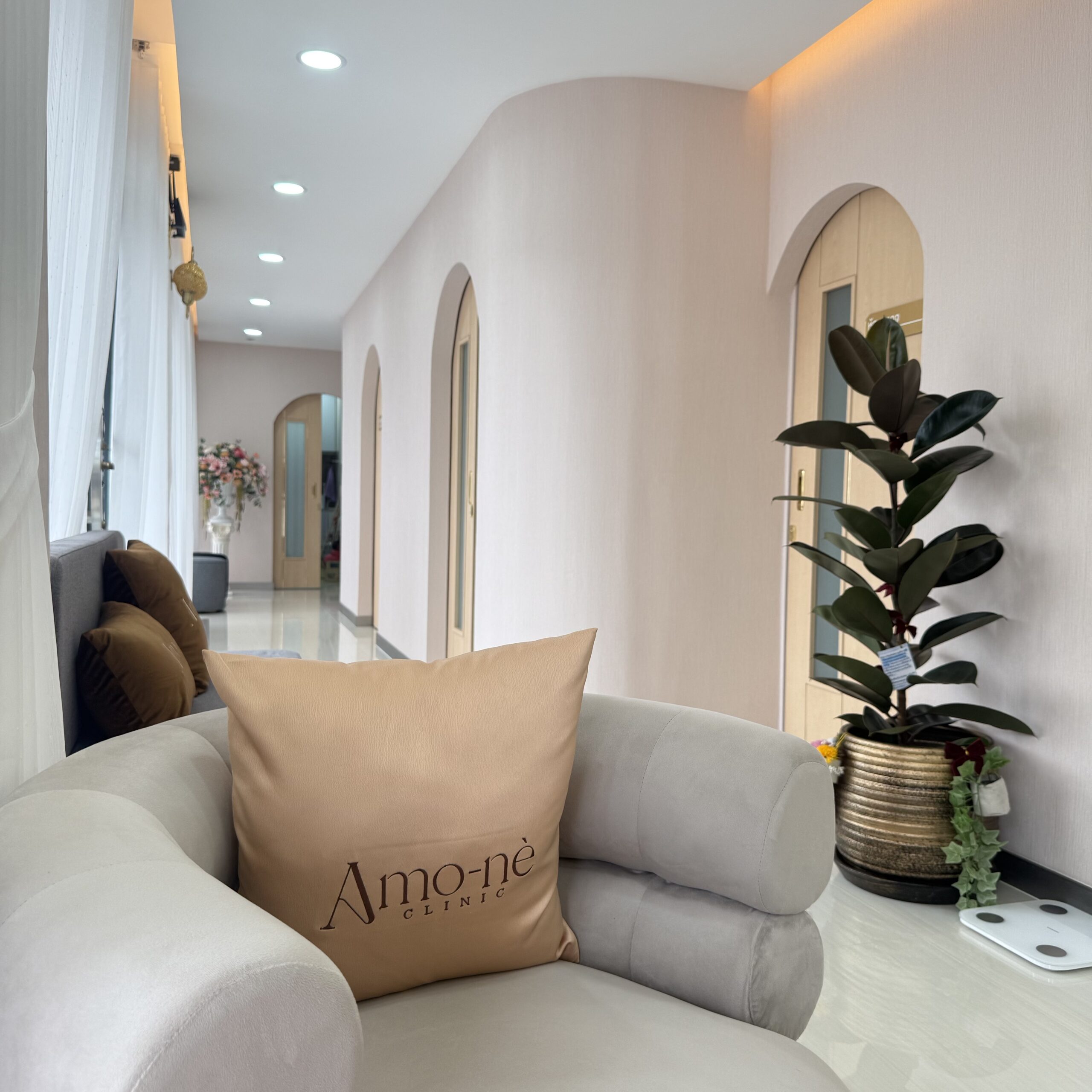 โซฟาสีขาวพร้อมหมอนแบรนด์ Amo-nè Clinic ในห้องรับรองตกแต่งสไตล์มินิมอลสีขาวพร้อมซุ้มโค้งและต้นไม้ประดับ