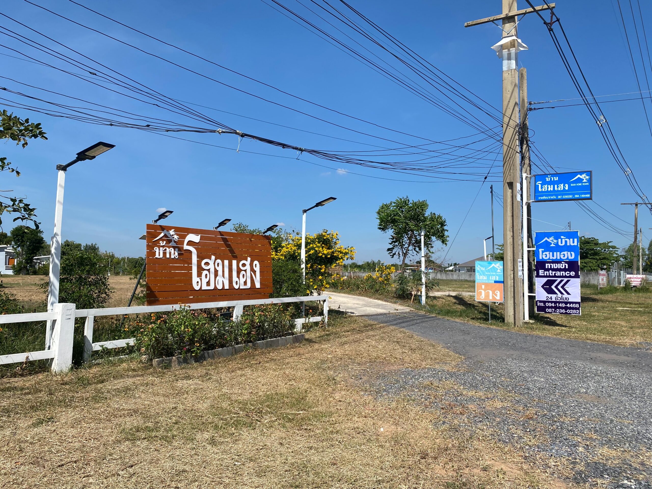 ป้ายทางเข้าบ้านโฮมเฮง พร้อมป้ายไม้สีน้ำตาลและป้ายสีน้ำเงินแสดงเบอร์โทรศัพท์และทางเข้า