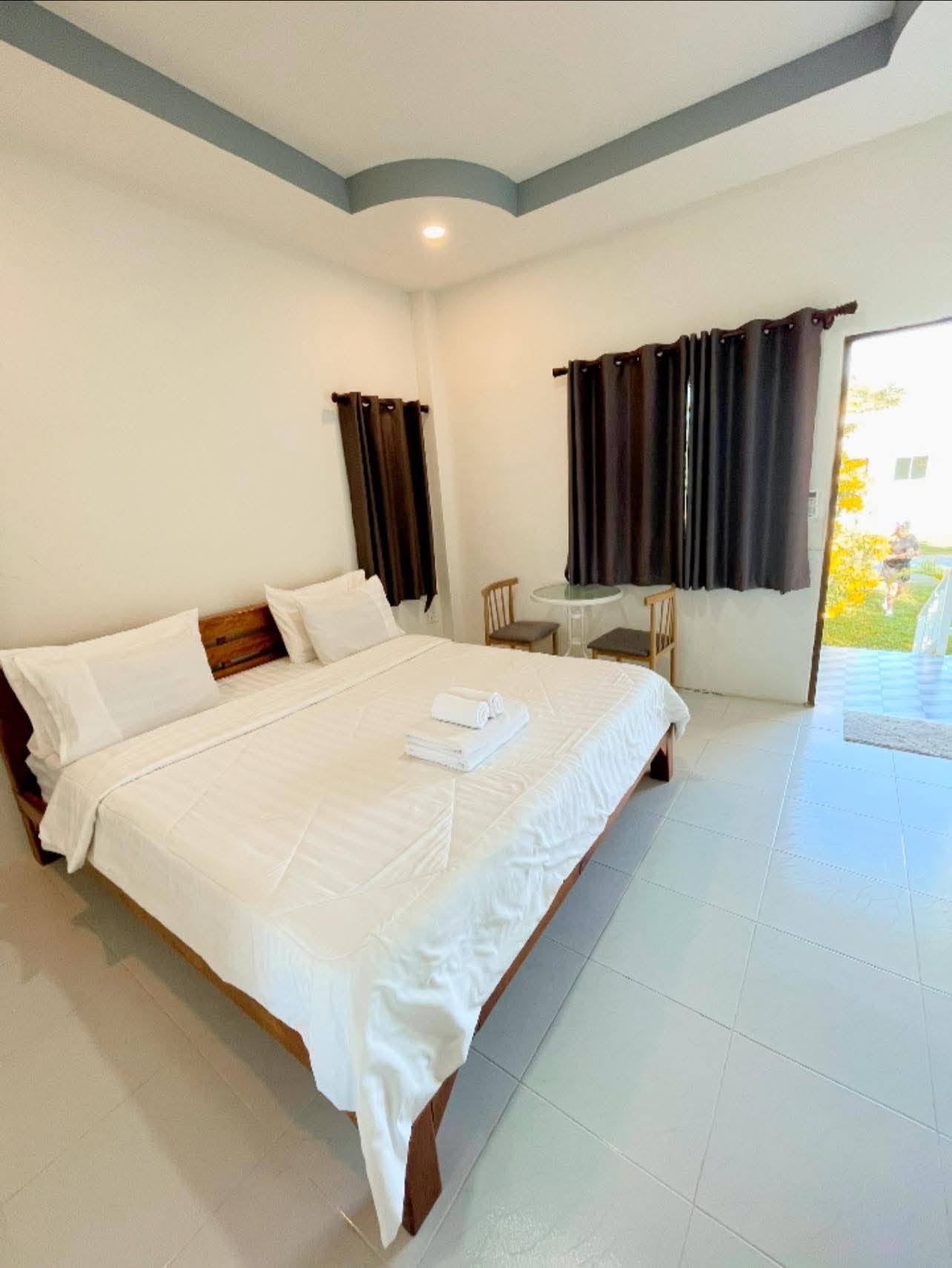 ห้องนอนโรงแรมสะอาดตกแต่งสีขาว มีเตียงคิงไซส์พร้อมผ้าปูสีขาว ผ้าม่านสีเข้ม และประตูเปิดออกสู่สวนสีเขียว