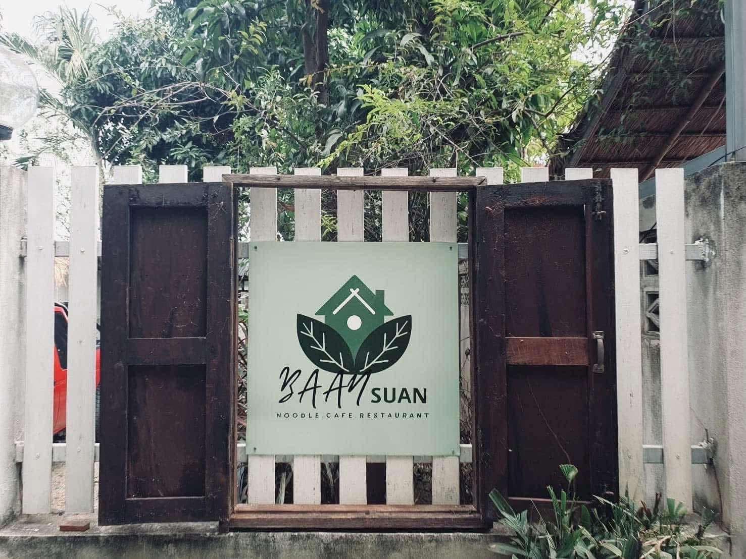 ป้ายร้านบ้านสวน นู้ดเดิล คาเฟ่ เรสเตอรองท์ ติดรั้วไม้สีขาวพร้อมหน้าต่างไม้เก่าและโลโก้รูปบ้านกับใบไม้สีเขียว