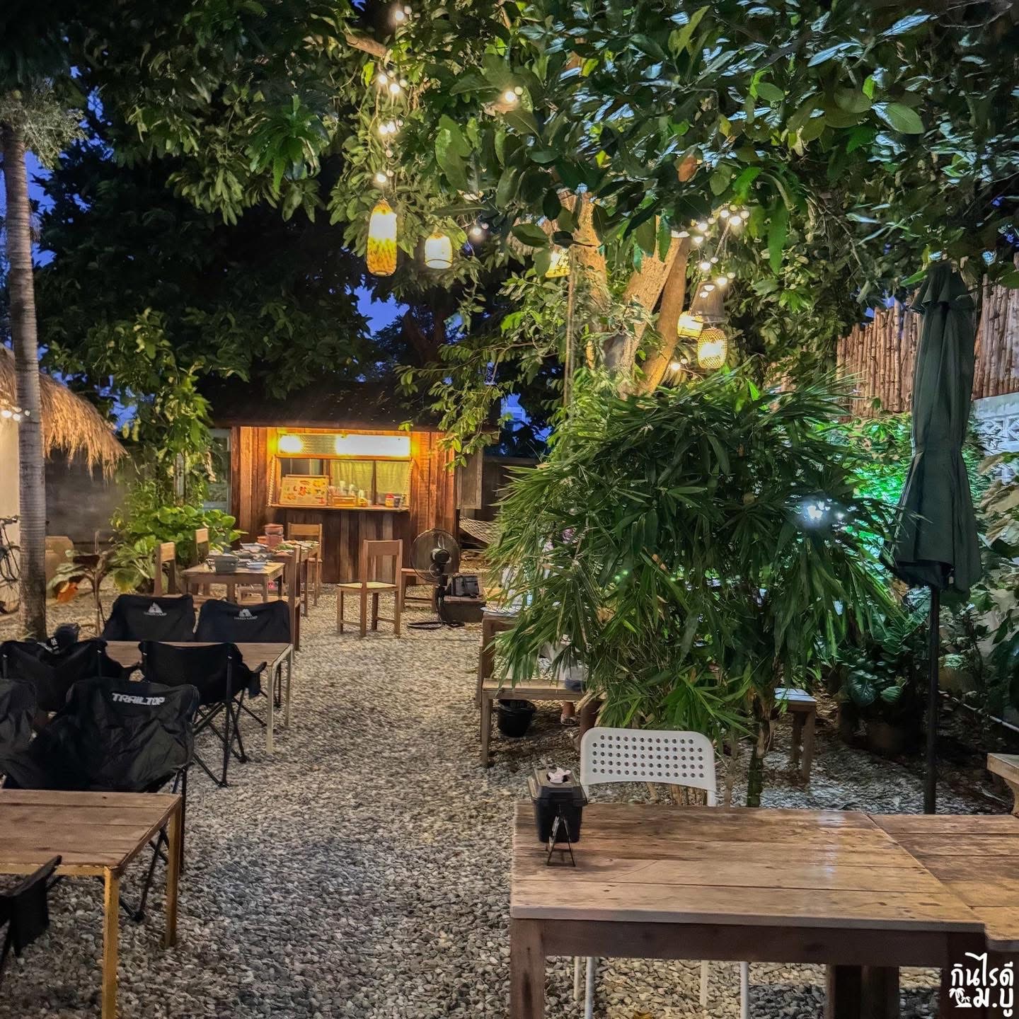 บรรยากาศร้านอาหารสวนกลางแจ้งยามเย็น มีโต๊ะไม้ เก้าอี้แคมป์ ไฟประดับบนต้นไม้ใหญ่ และซุ้มขายเครื่องดื่มไม้สไตล์คาเฟ่