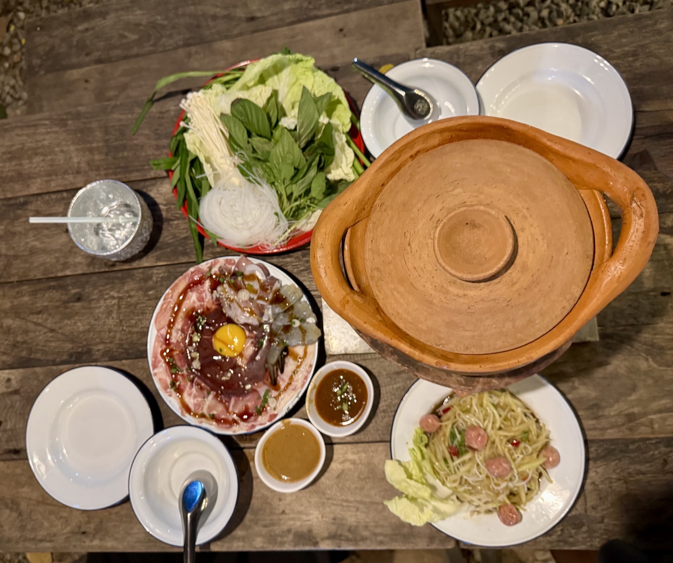 ชุดจิ้มจุ่มหม้อดินพร้อมผักสด เนื้อสัตว์หมูสไลด์กุ้งไข่แดง น้ำจิ้ม และส้มตำแหนมบนโต๊ะไม้