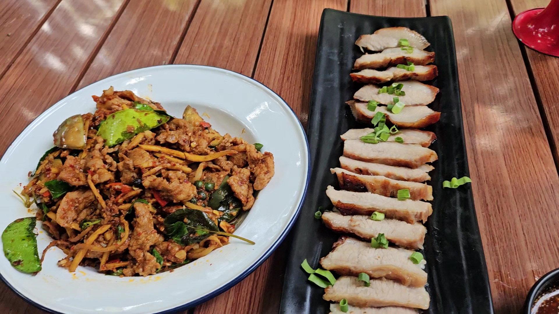 ผัดฉ่าหมูใส่สมุนไพรจานขาวคู่กับหมูอบสไลด์เรียงบนจานดำโรยต้นหอม บนโต๊ะไม้