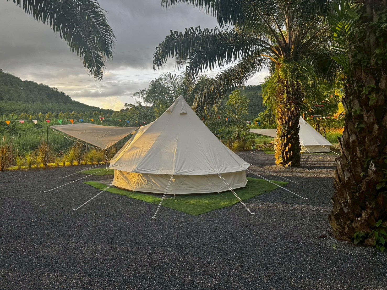 เต็นท์กระโจมสีขาวสไตล์แกลมปิ้งบนพื้นหญ้าเทียมสีเขียว ท่ามกลางต้นปาล์มและธงสามเหลี่ยมหลากสี บรรยากาศภูเขาและท้องฟ้าครึ้มฝน