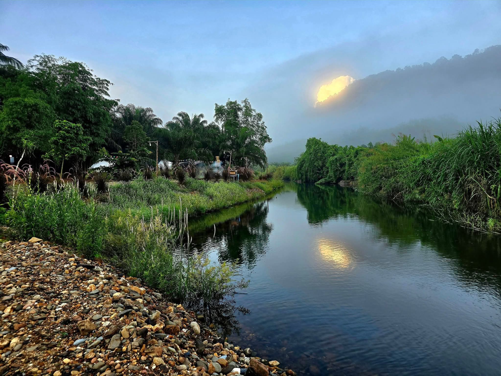 วิวแม่น้ำยามเช้าท่ามกลางหมอกและต้นไม้เขียวขจี พร้อมแสงแดดส่องผ่านเมฆเหนือภูเขา