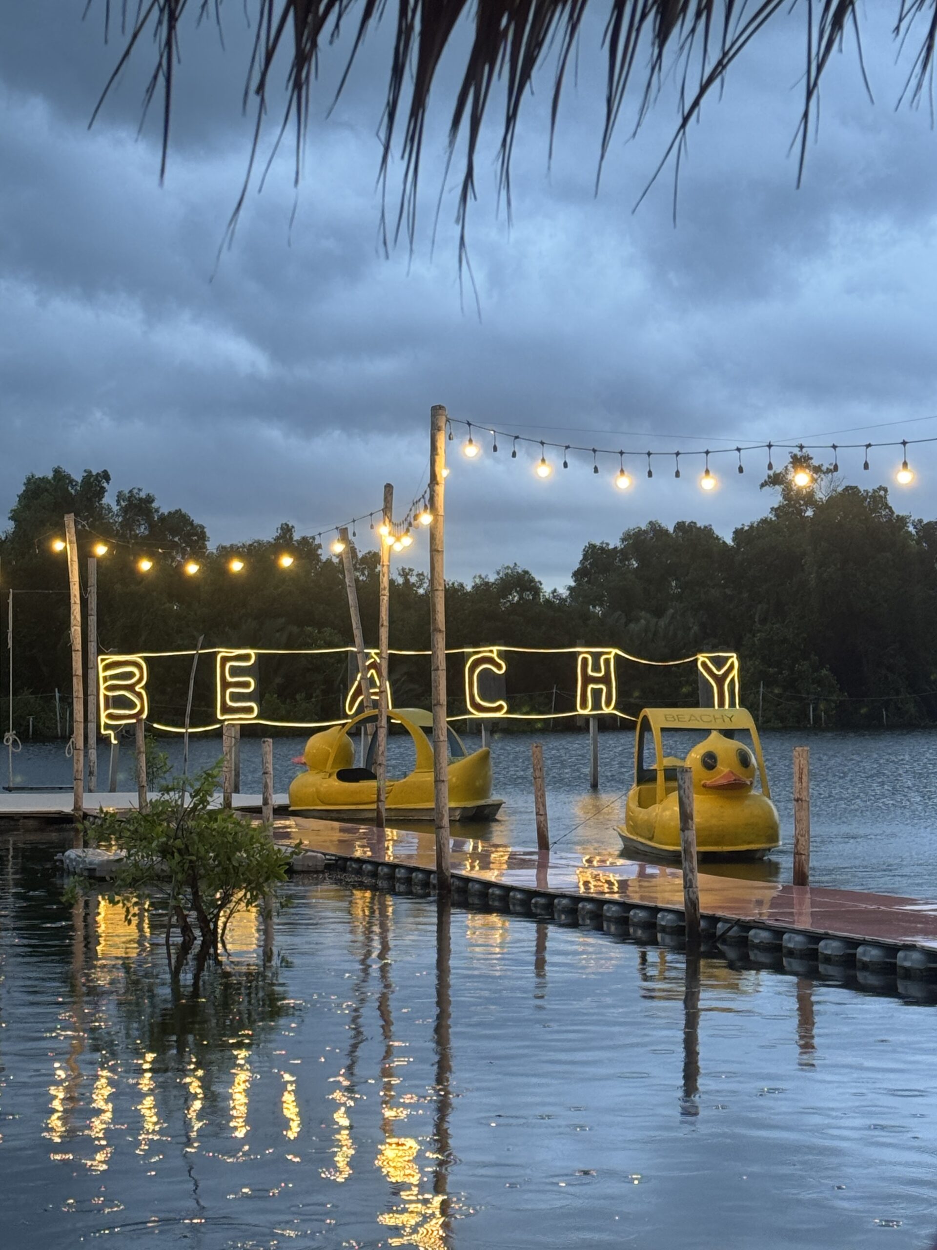 ท่าเรือริมน้ำร้าน Beachy ยามเย็น มีป้ายไฟนีออนสีเหลืองเขียนว่า BEACHY เรือปั่นรูปเป็ดสีเหลืองจอดอยู่บนท่า มีไฟสายประดับและบรรยากาศโรแมนติก