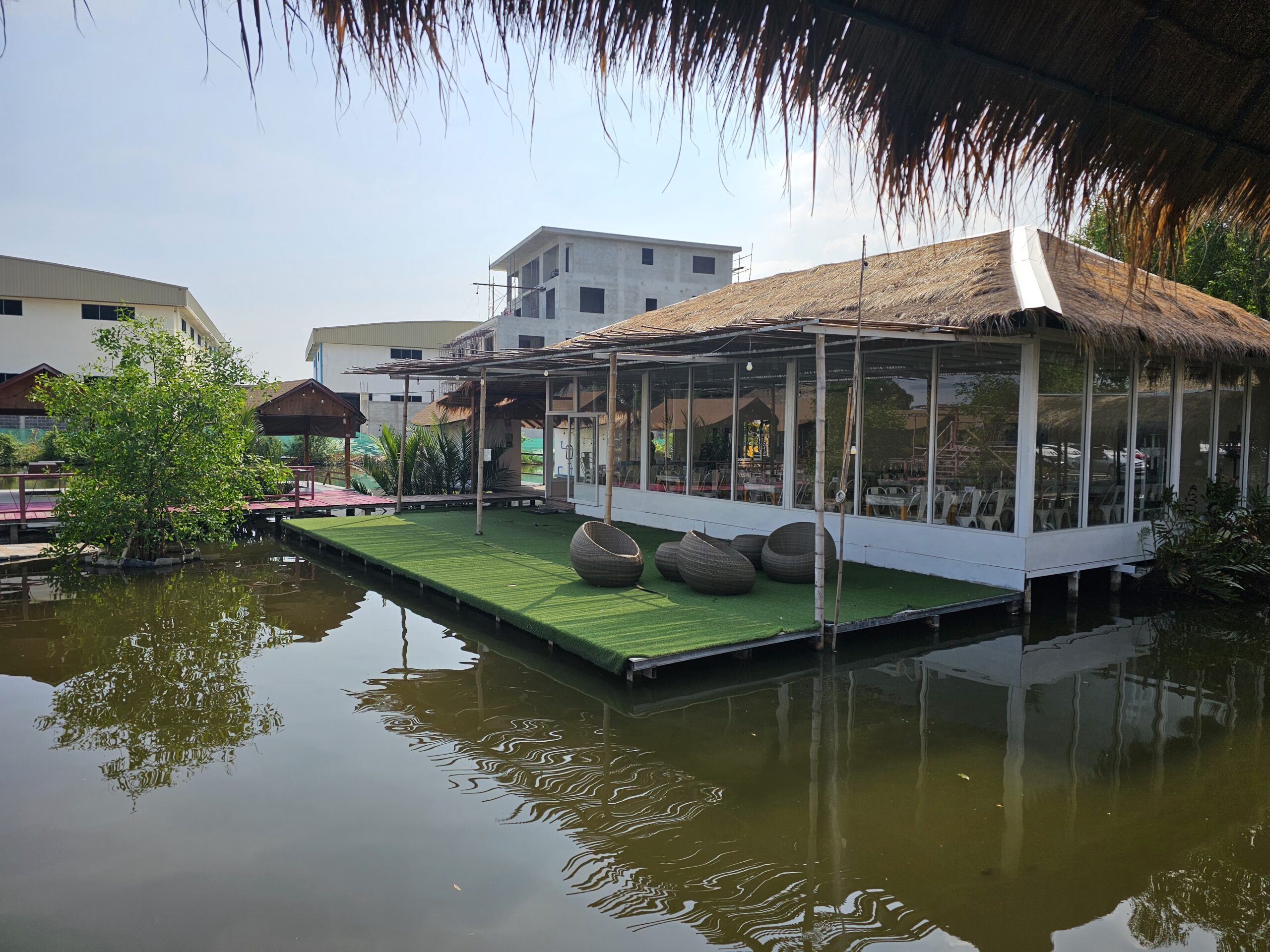 ร้านอาหารลอยน้ำหลังคามุงจากริมบ่อน้ำ มีระเบียงปูหญ้าเทียมสีเขียวและเก้าอี้หวายทรงกลม