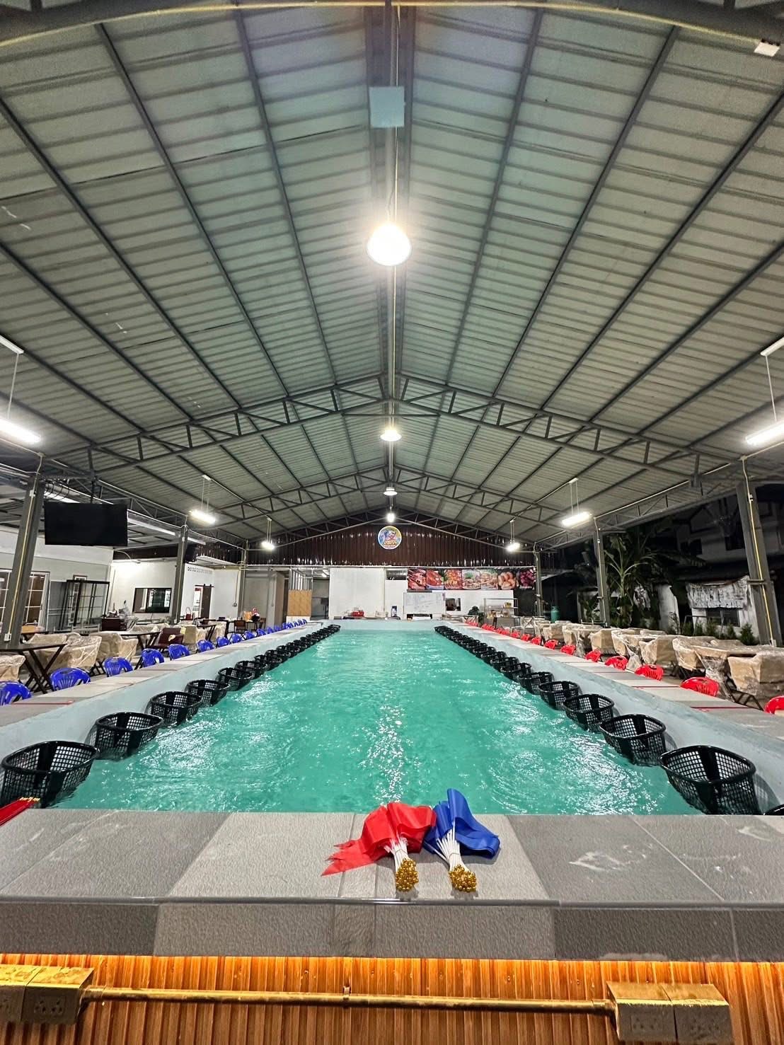 บ่อตกกุ้งในร่มขนาดใหญ่ภายใต้หลังคาโครงเหล็ก มีตะกร้าดำเรียงรายรอบบ่อน้ำสีเขียว พร้อมโต๊ะเก้าอี้สีน้ำเงินสำหรับลูกค้า