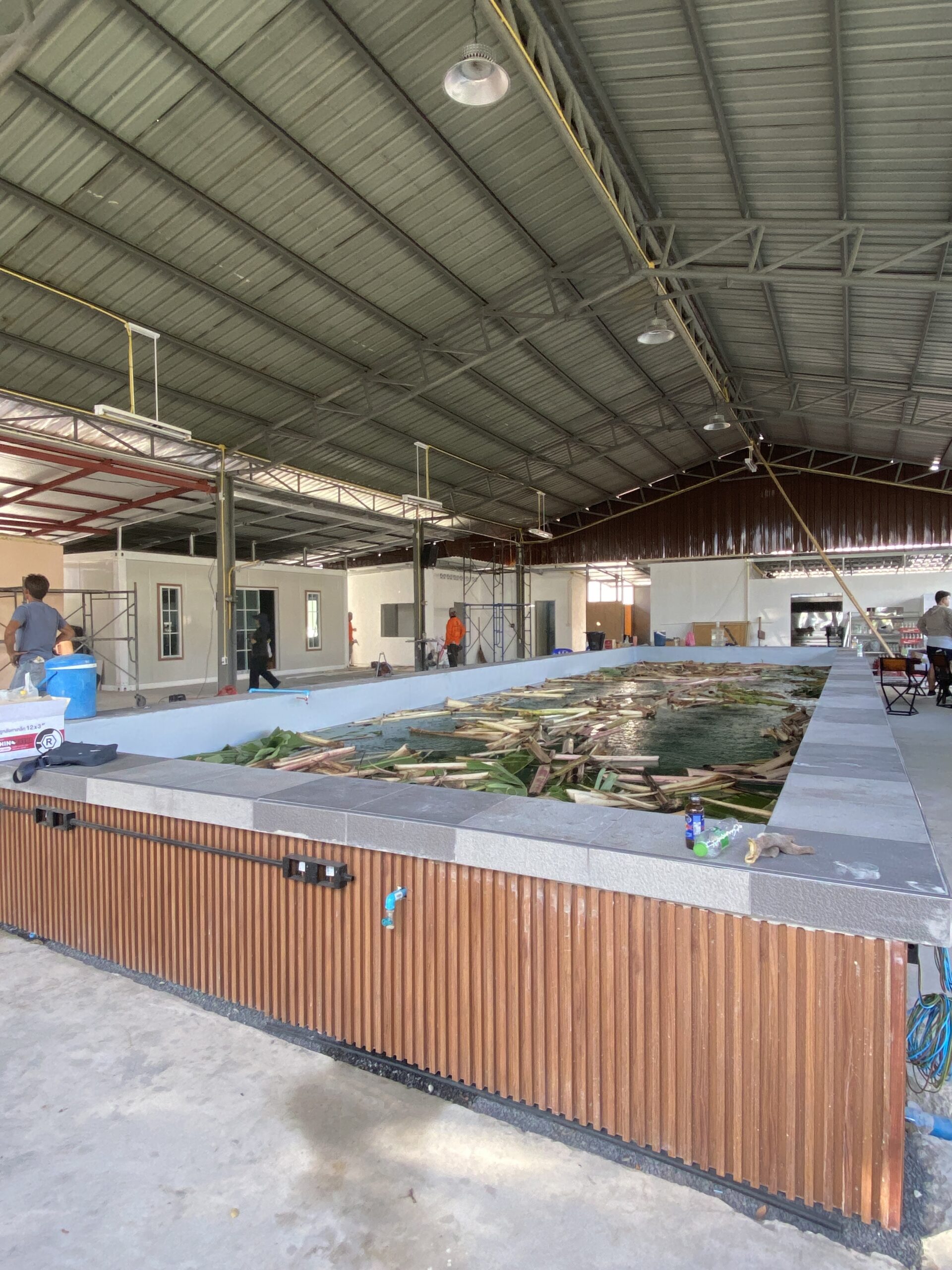 ภายในร้านอาหารที่กำลังก่อสร้าง มีบ่อน้ำขนาดใหญ่ตกแต่งด้วยไม้ระแนง อยู่ใต้หลังคาเหล็กขนาดใหญ่ มีนั่งร้านและคนงานกำลังทำงาน