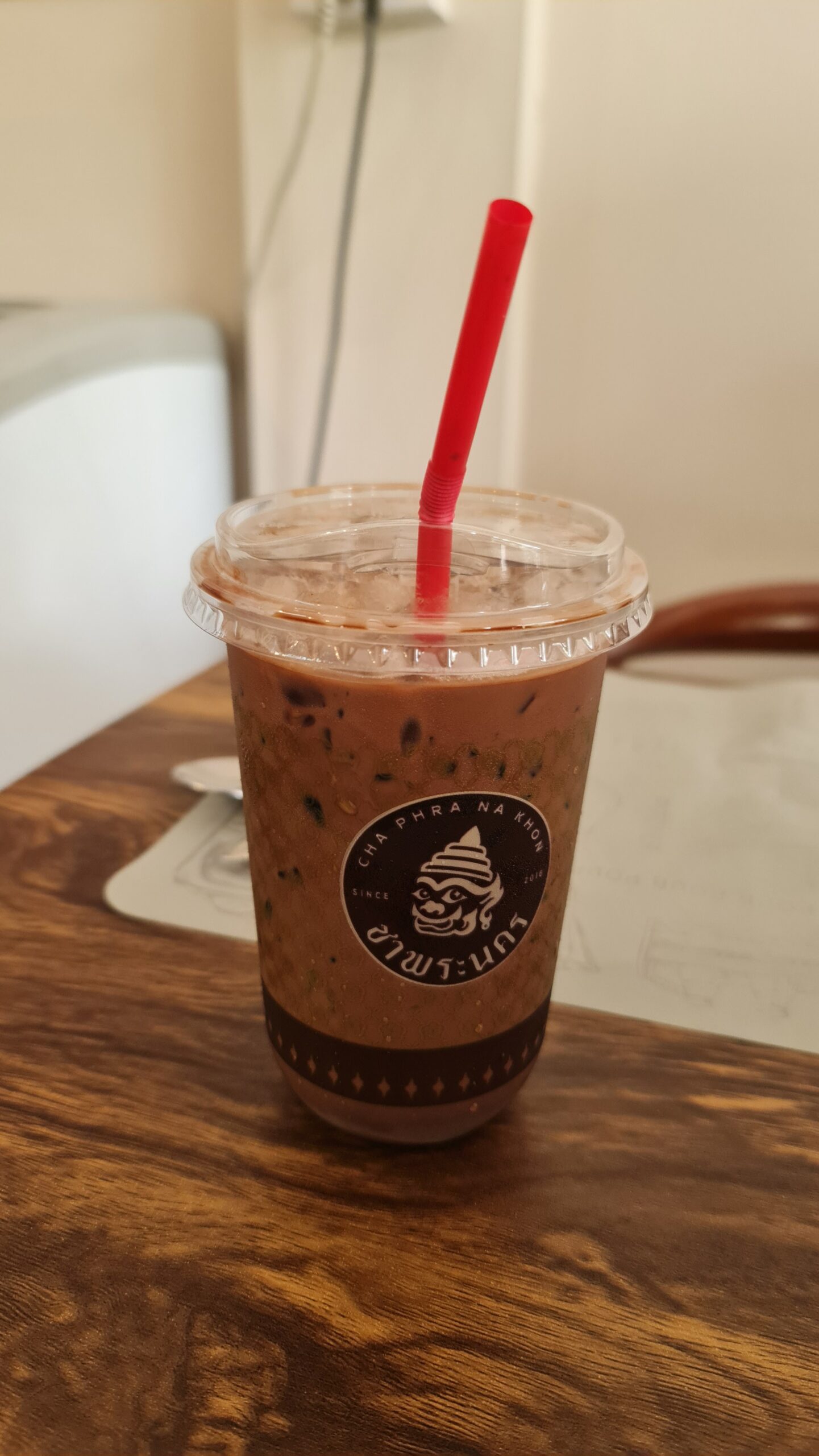 เครื่องดื่มช็อกโกแลตเย็นในแก้วพลาสติกของร้านชาพระนคร พร้อมหลอดสีแดง วางบนโต๊ะไม้