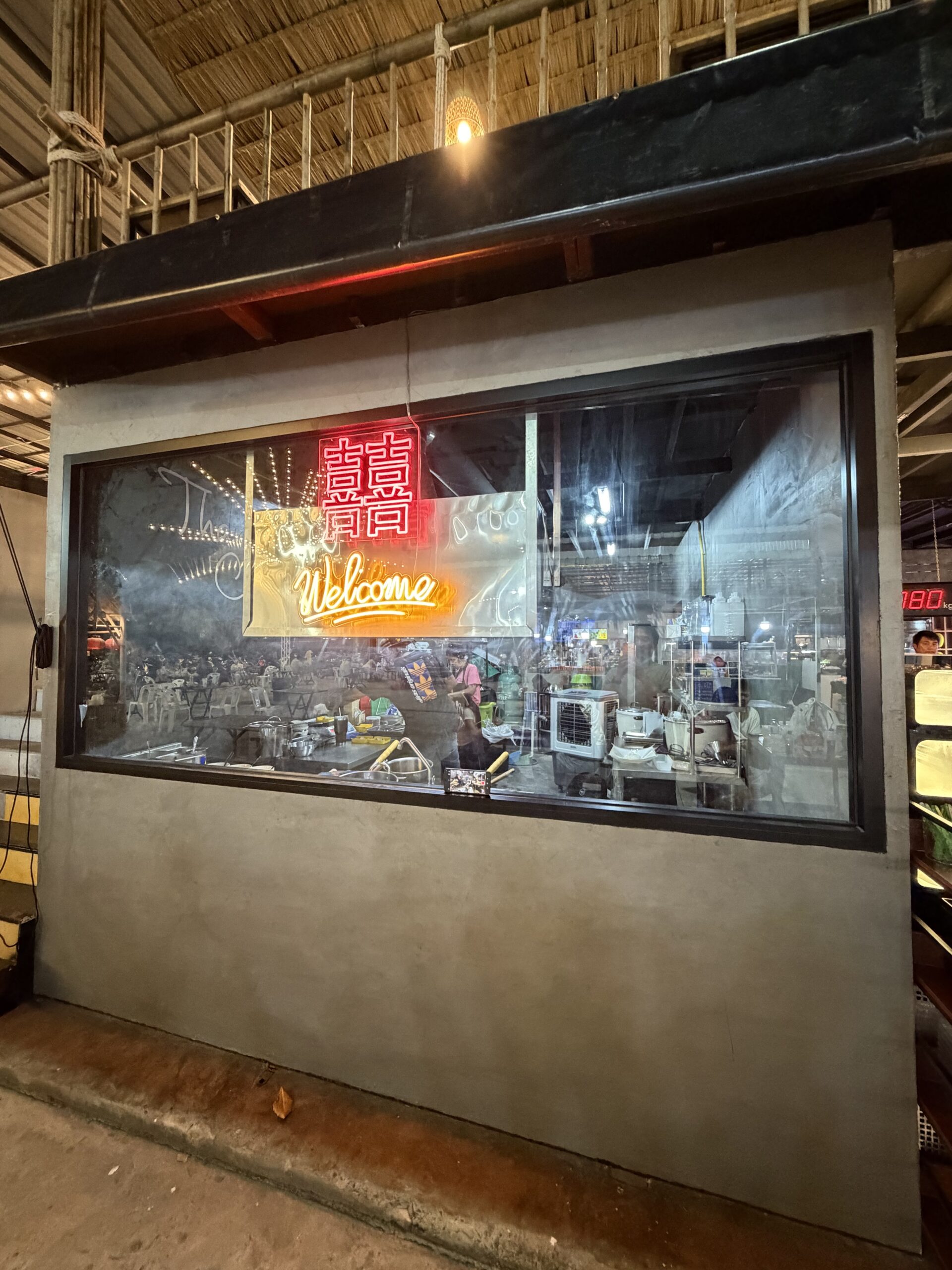 หน้าต่างกระจกร้านอาหารจีนที่มีป้ายไฟนีออนสีแดงตัวอักษรจีน囍และข้อความ Welcome สีเหลือง มองเห็นครัวและบรรยากาศภายในร้าน