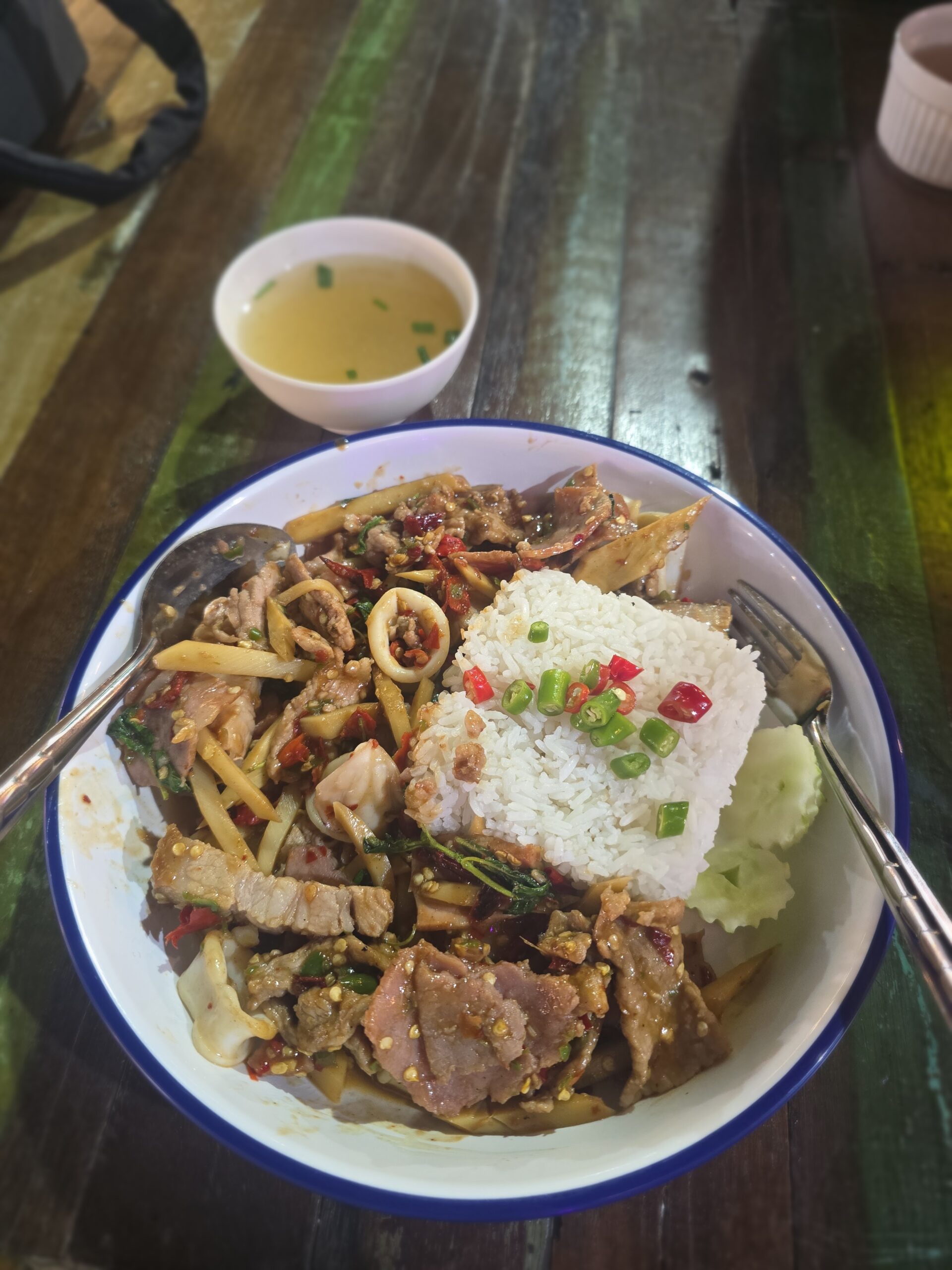 จานข้าวราดผัดเผ็ดหมูและปลาหมึกกับหน่อไม้ พริก โหระพา เสิร์ฟพร้อมข้าวสวย แตงกวา และน้ำซุป บนโต๊ะไม้