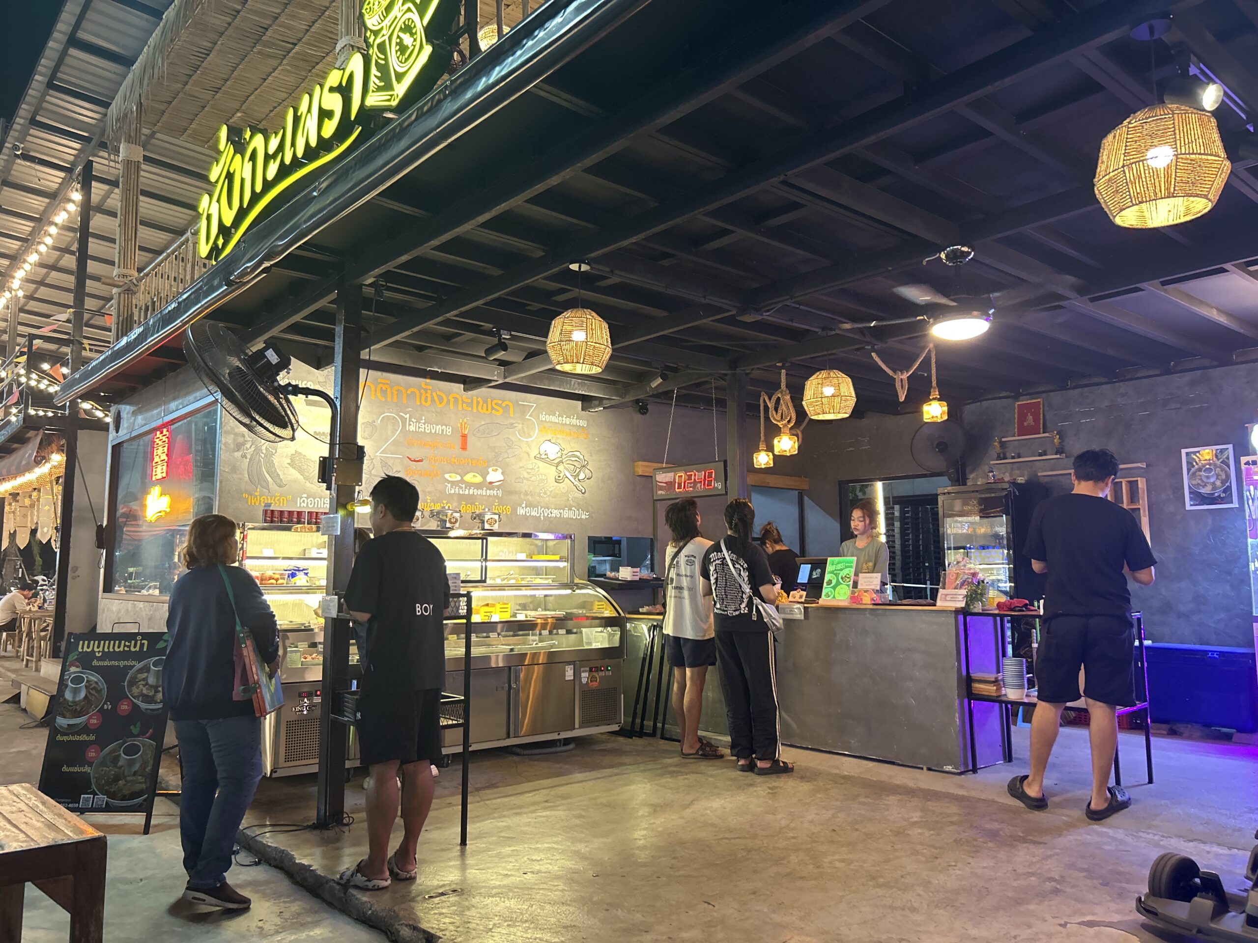 ร้านอาหารไทยสไตล์สตรีทฟู้ดที่มีป้ายไฟสีเขียวเขียนว่าชูใจกะเพรา มีลูกค้ายืนต่อคิวสั่งอาหารหน้าตู้โชว์อาหาร โคมไฟหวายห้อยจากเพดาน บรรยากาศร้านแบบตลาดกลางคืน