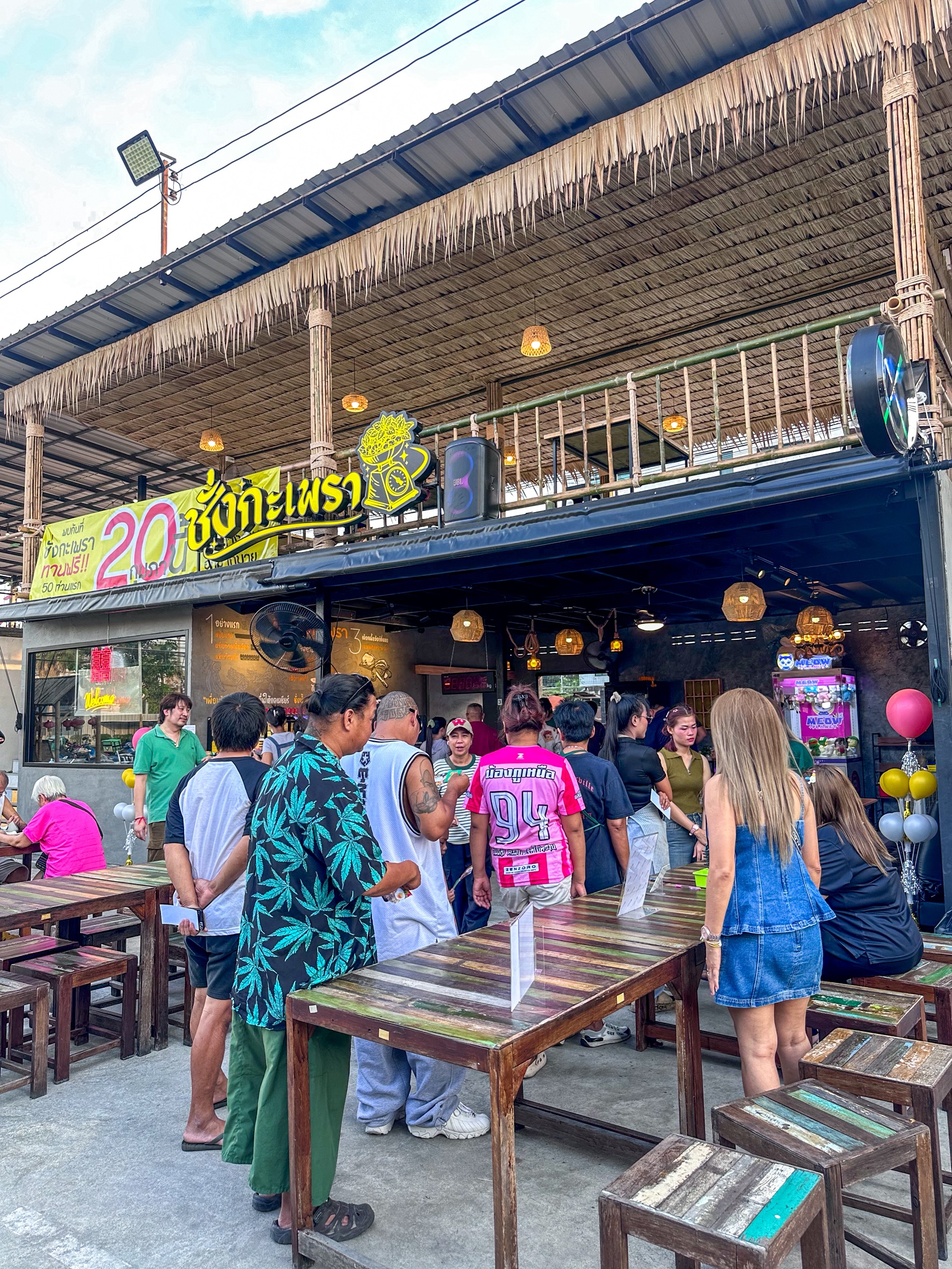 ร้านอาหารไทยชื่อ ชั่งกะเพรา บรรยากาศคึกคัก มีลูกค้ายืนรอคิวหน้าร้าน มีโต๊ะไม้สีสันและหลังคามุงจาก ป้ายโปรโมชั่น 20 บาท