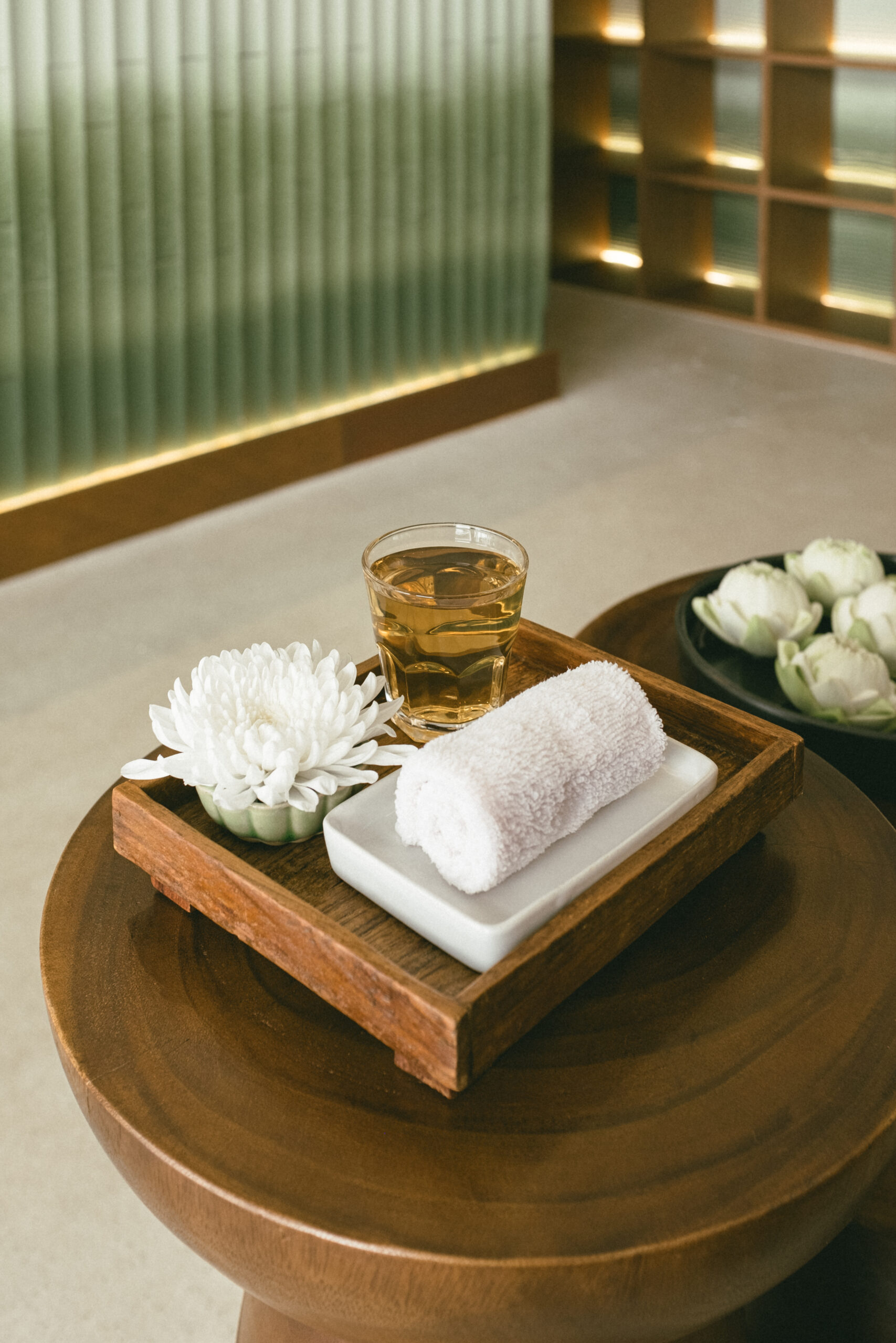 ถาดไม้บนโต๊ะกลมวางชาเย็นในแก้วใส ผ้าเช็ดหน้าม้วนสีขาว และดอกเบญจมาศขาวในบรรยากาศสปาหรูหรา