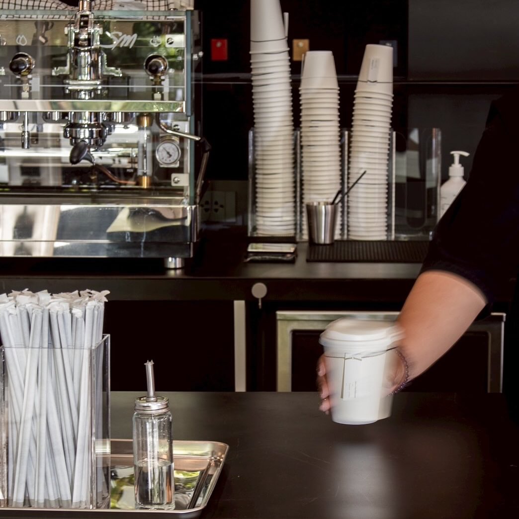 บาริสต้ากำลังเสิร์ฟกาแฟในแก้วกระดาษสีขาวบนเคาน์เตอร์ร้านกาแฟพร้อมเครื่องชงเอสเปรสโซ
