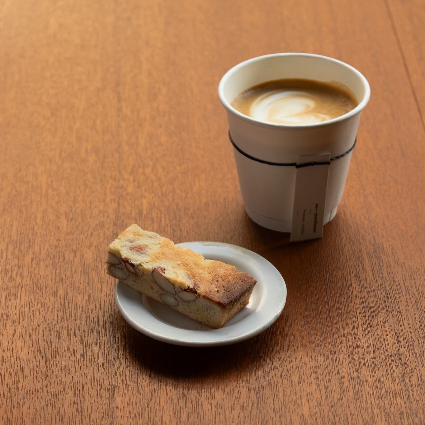 กาแฟลาเต้ในแก้วกระดาษพร้อมบิสคอตตี้อัลมอนด์บนจานเล็กวางบนโต๊ะไม้