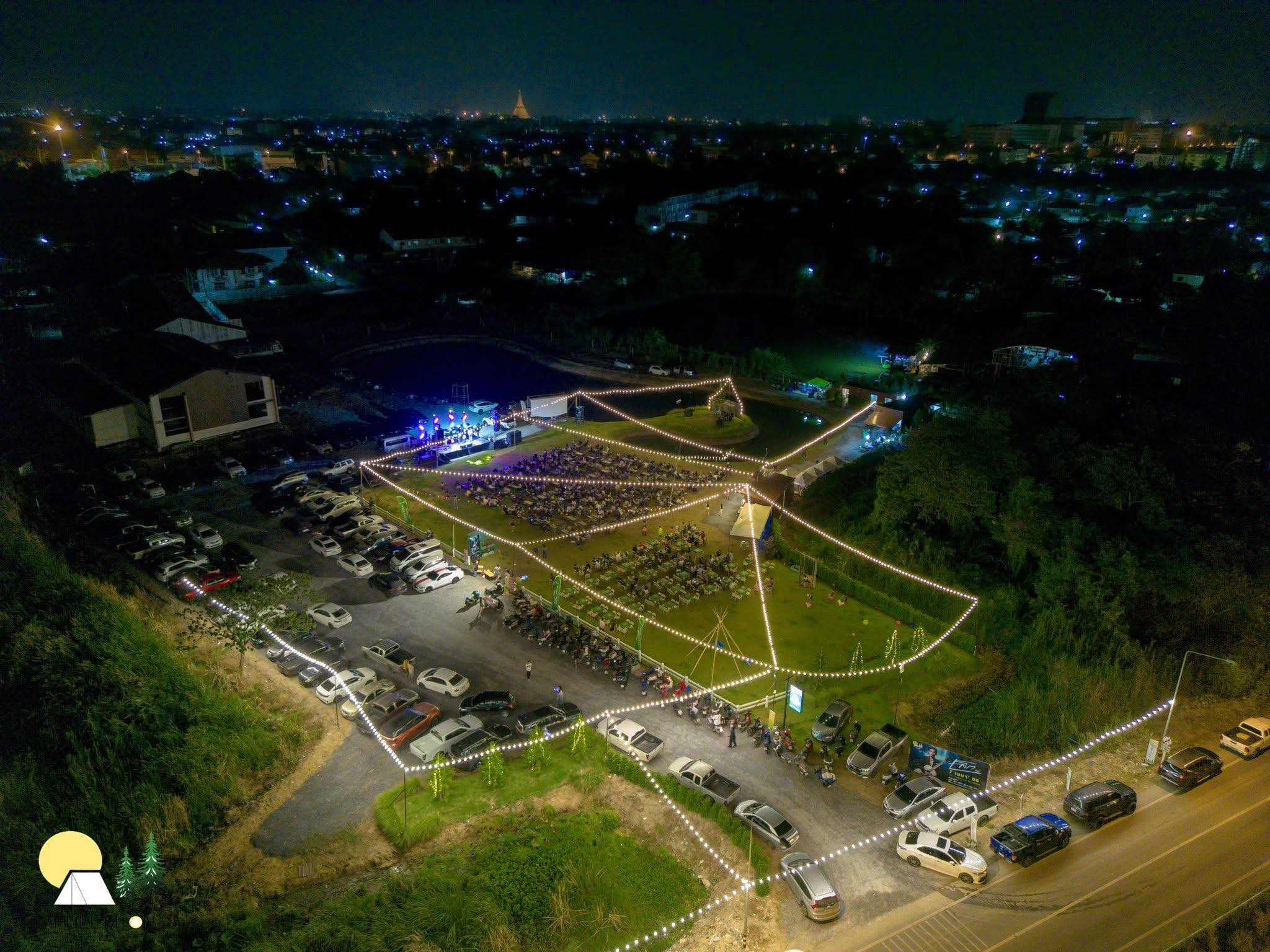 ภาพมุมสูงของสถานที่จัดงานกลางแจ้งในเวลากลางคืน มีไฟประดับสายสว่างไสว เวทีคอนเสิร์ต ลานจอดรถ และผู้คนนั่งชมการแสดงบนสนามหญ้า โดยมีวิวเมืองเป็นฉากหลัง