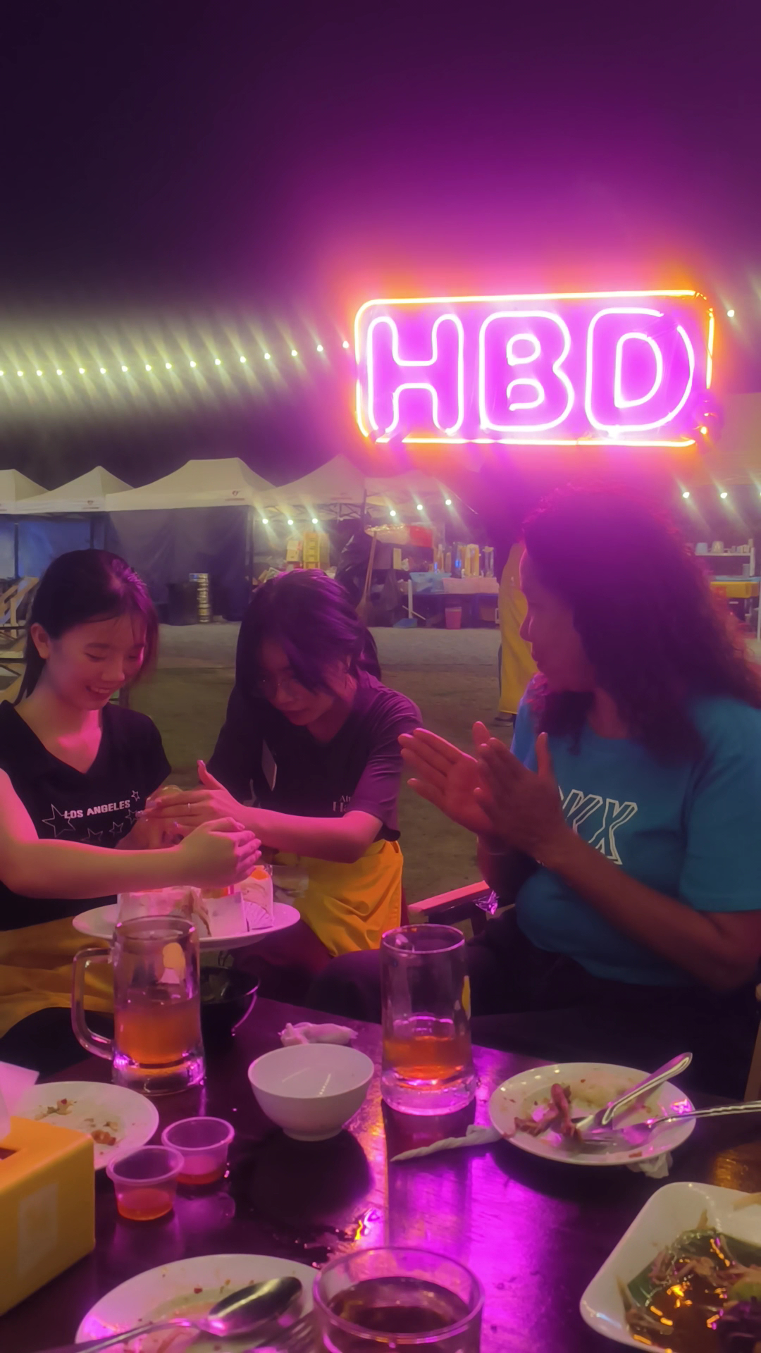 กลุ่มเพื่อนสาวนั่งฉลองวันเกิดที่ร้านอาหารกลางแจ้งใต้ป้ายไฟนีออน HBD สีชมพูม่วง พร้อมเครื่องดื่มและอาหารบนโต๊ะ