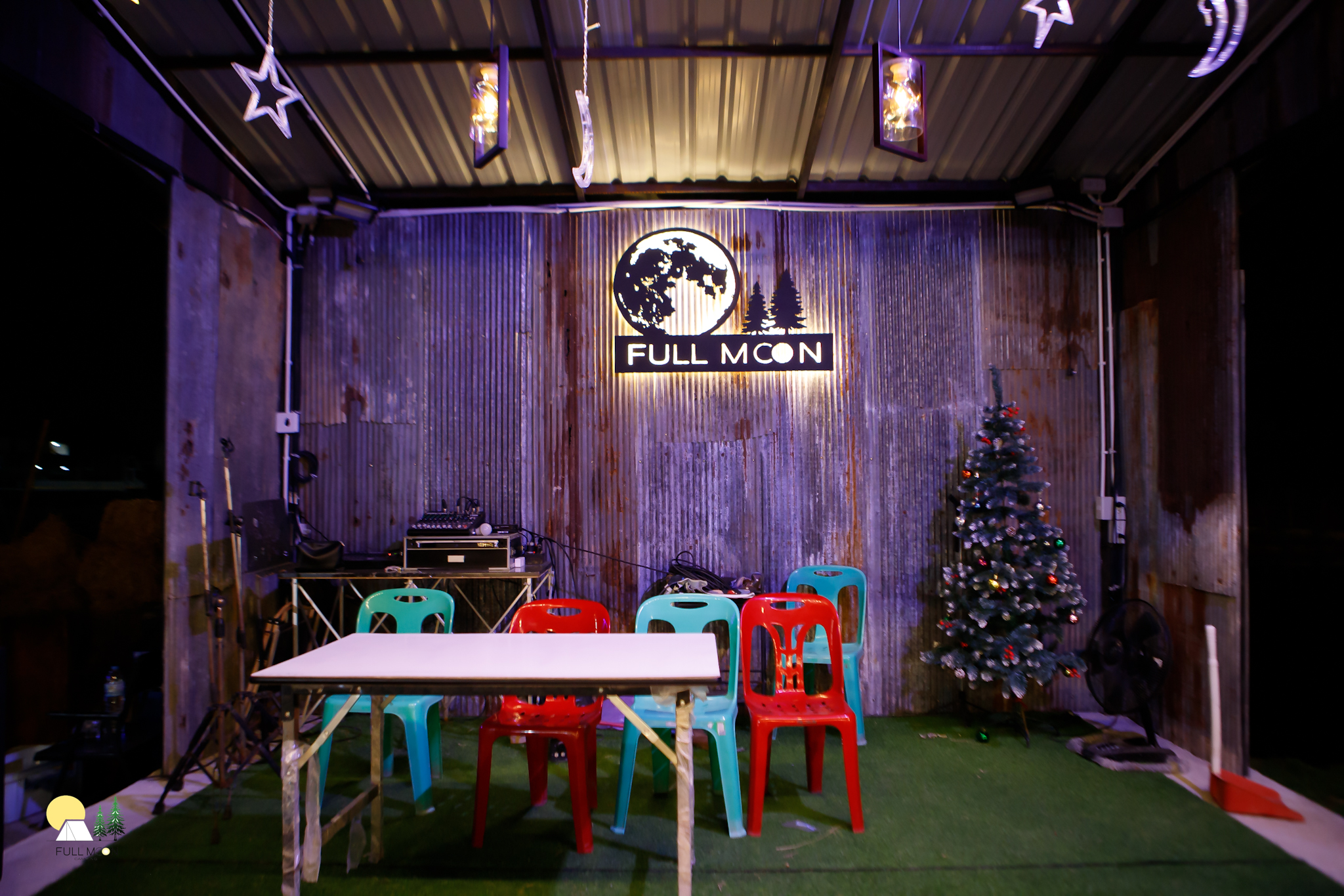 พื้นที่นั่งเล่นของร้าน Full Moon ตกแต่งสไตล์ลอฟท์ผนังสังกะสี มีเก้าอี้พลาสติกสีเขียวและแดง โต๊ะพับ ต้นคริสต์มาส และเวทีดีเจพร้อมอุปกรณ์เครื่องเสียง