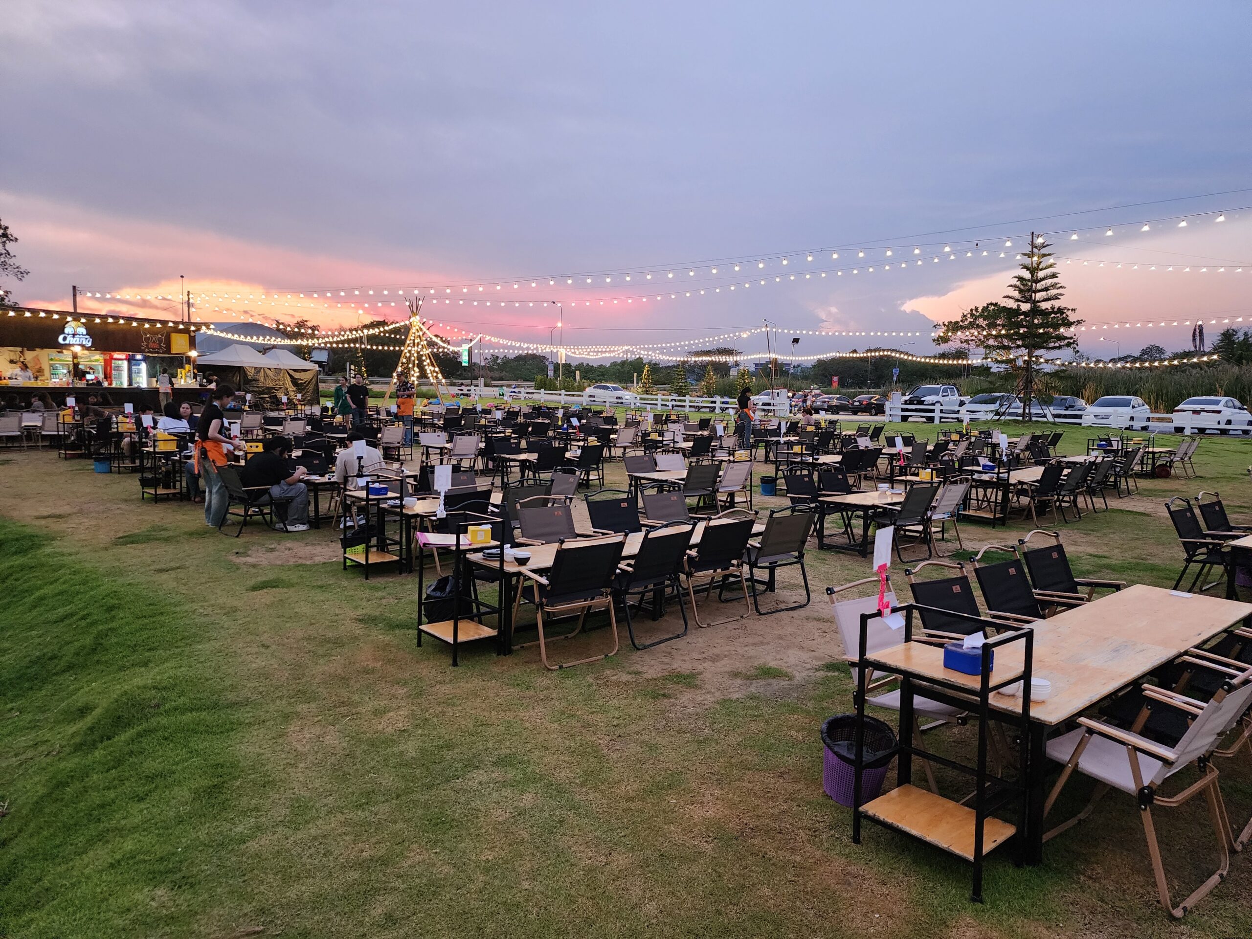 ร้านอาหารกลางแจ้งบนสนามหญ้าสีเขียวพร้อมโต๊ะเก้าอี้แคมป์ปิ้งจำนวนมาก ประดับไฟสายระยิบระยับยามพระอาทิตย์ตกดิน มีป้ายเบียร์ช้างและเต็นท์ทีปี