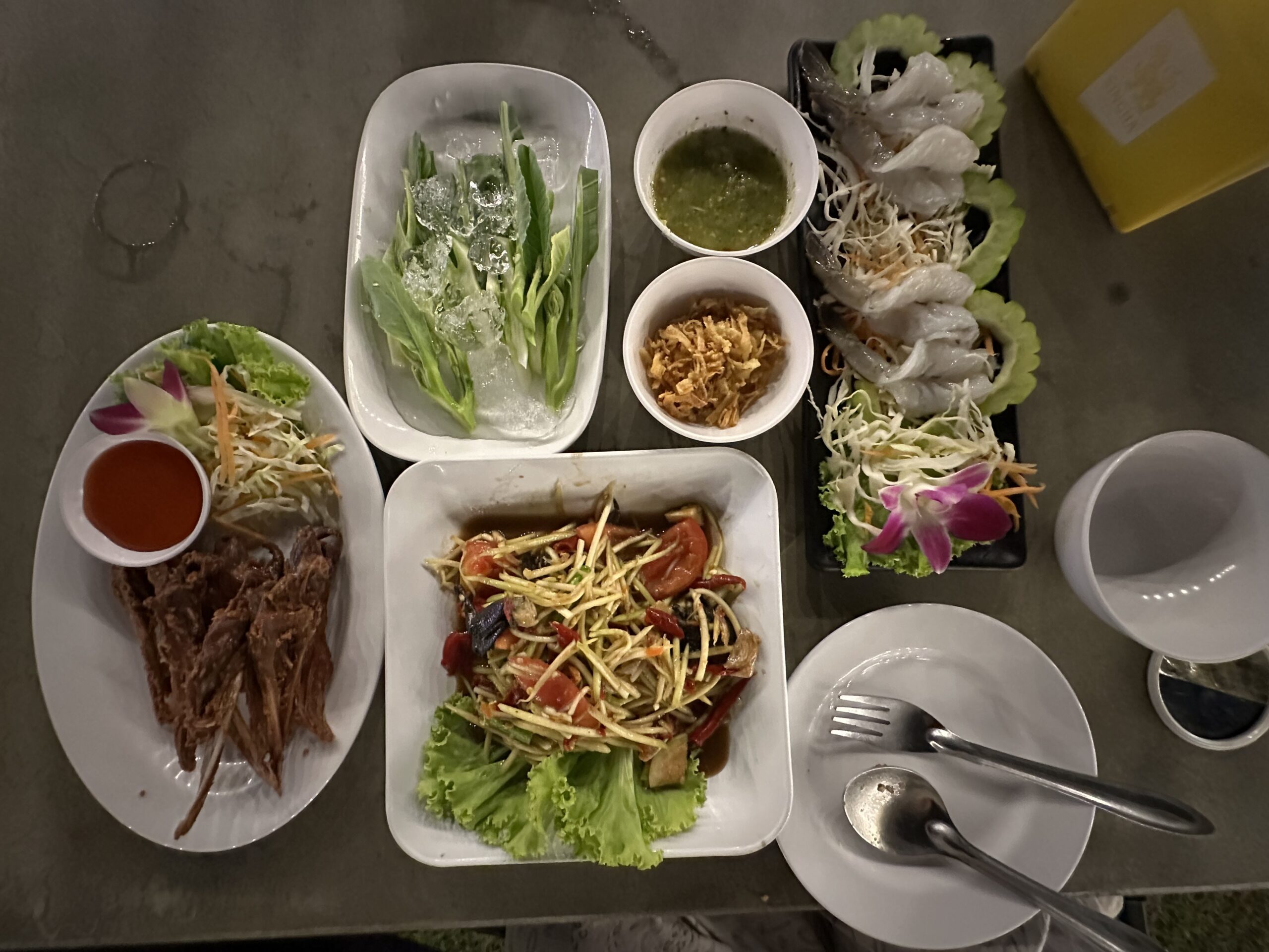 อาหารไทยหลายจานบนโต๊ะ ได้แก่ ส้มตำ กุ้งแช่น้ำปลา ผักสดแช่น้ำแข็ง เนื้อทอด และน้ำจิ้มซีฟู้ด