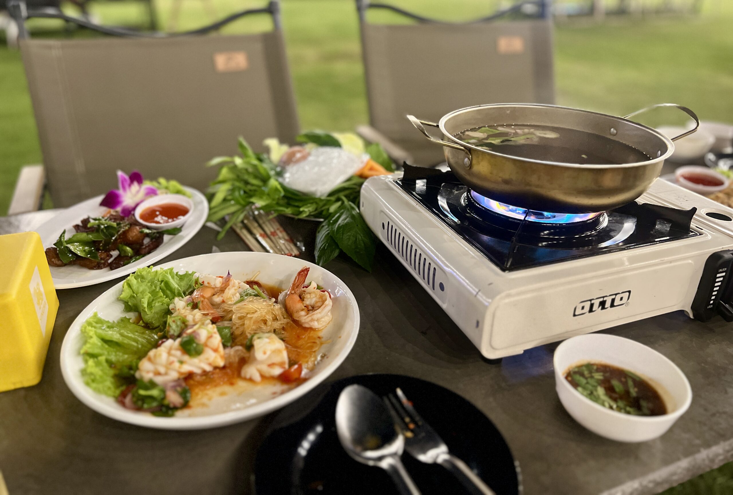 โต๊ะอาหารกลางแจ้งพร้อมเตาแก๊สพกหา OTTO หม้อสุกี้ จานกุ้งสดยำวุ้นเส้น และจานเนื้อทอดกระเทียมพริกไทย บนพื้นหลังสนามหญ้าเขียวขจี