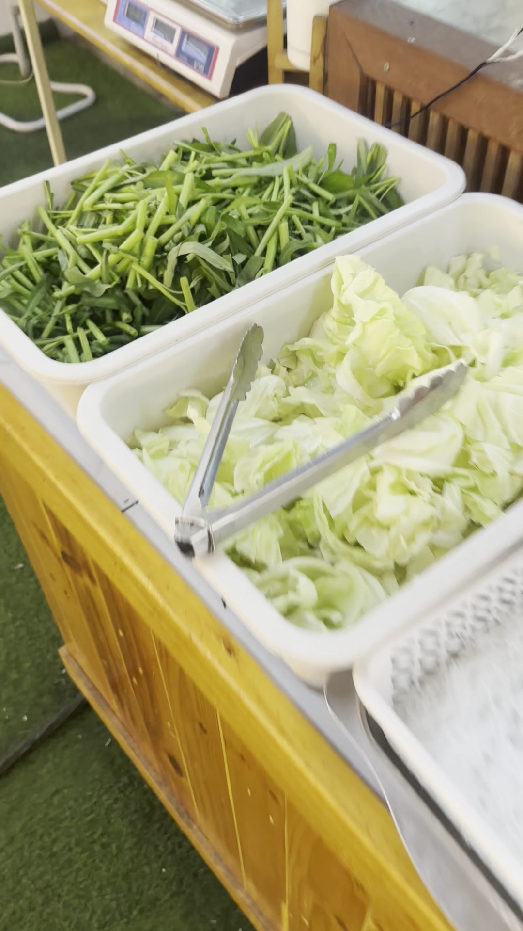 บุฟเฟ่ต์ผักสด มีผักบุ้งและกะหล่ำปลีหั่นในถาดขาว พร้อมที่คีบ สำหรับชาบูหรือหม้อไฟ