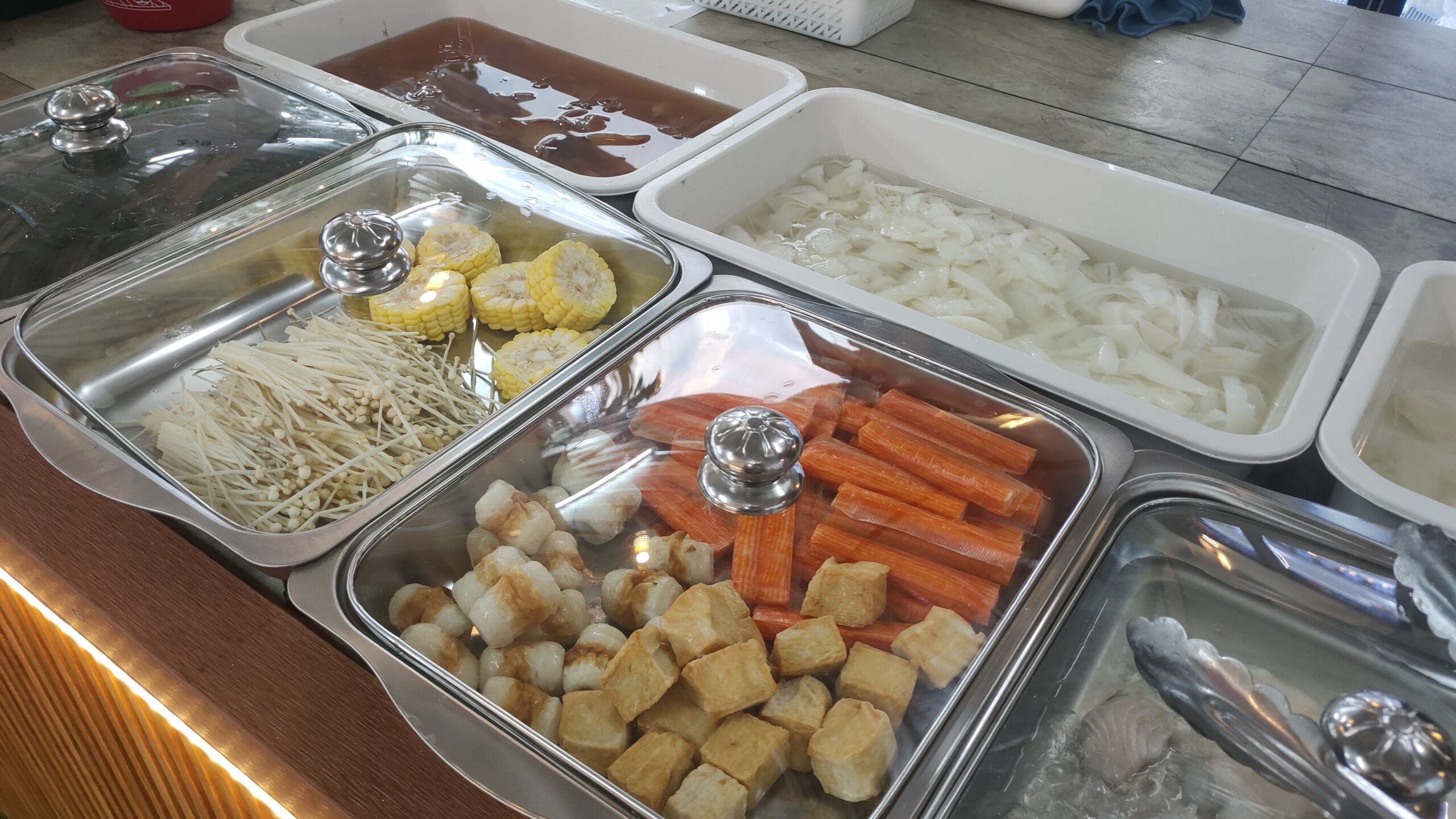 บุฟเฟ่ต์ชาบูพร้อมวัตถุดิบหลากหลาย เช่น เห็ดเข็มทอง ข้าวโพด เต้าหู้ทอด ปูอัด และเส้นก๋วยเตี๋ยวในถาดสแตนเลส