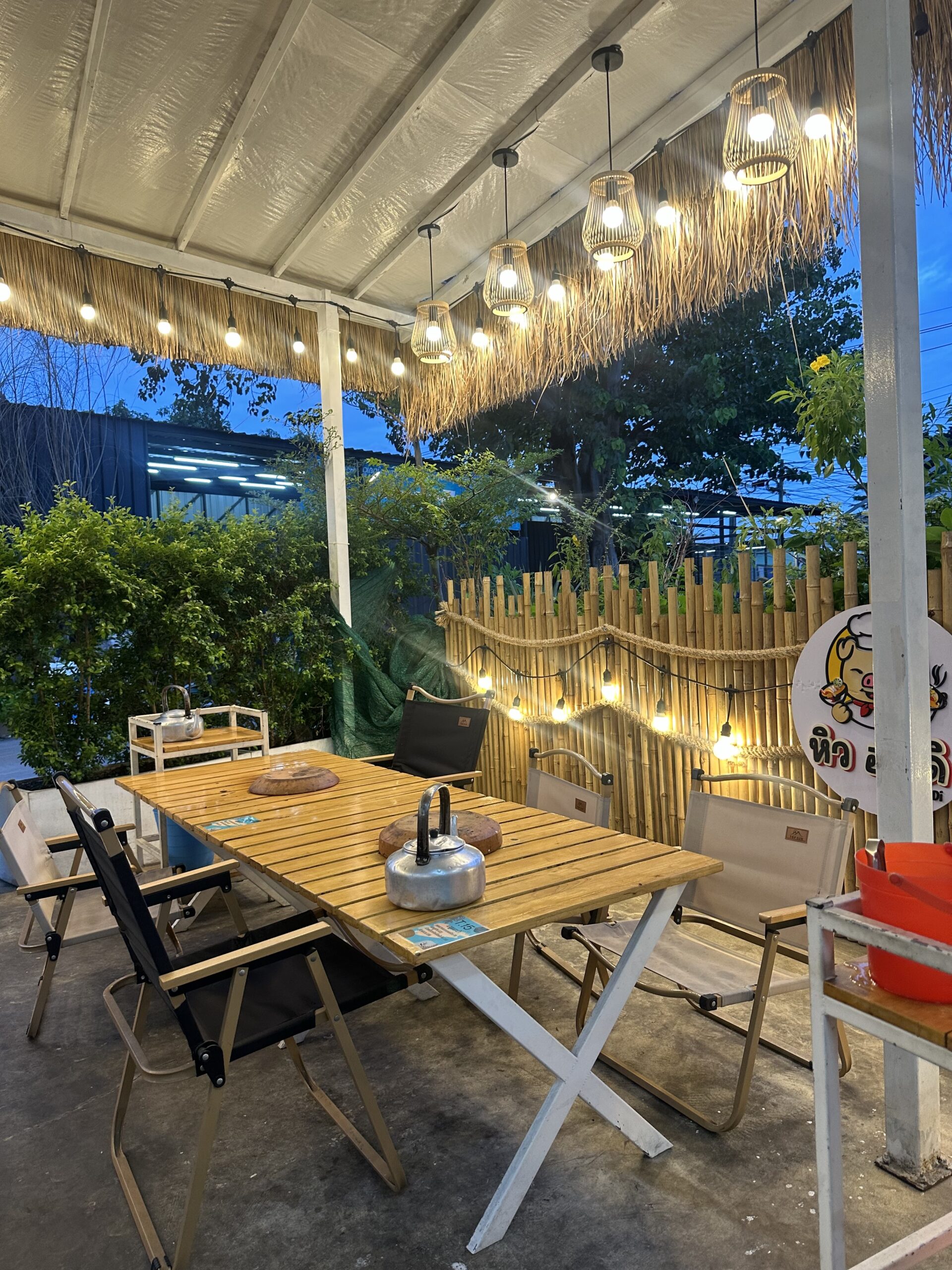 โซนนั่งกลางแจ้งของร้านอาหารตกแต่งด้วยรั้วไม้ไผ่ โคมไฟหวาย โต๊ะไม้พับพร้อมเก้าอี้แคมป์ปิ้ง บรรยากาศอบอุ่นยามเย็น