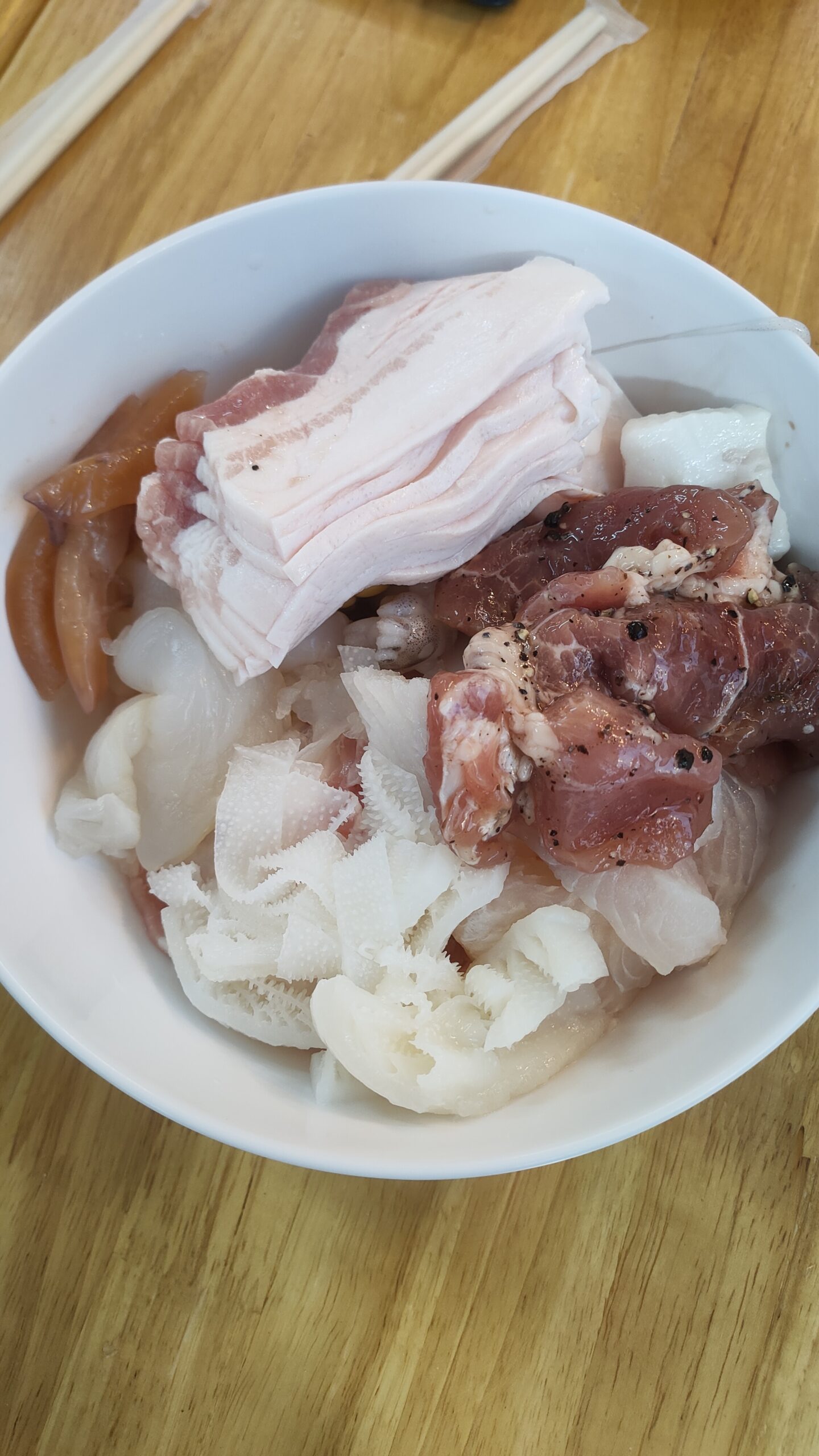 ชามเนื้อสดรวมสำหรับปิ้งย่างหรือชาบู มีหมูสามชั้น เนื้อหมักพริกไทย เครื่องในและหมึกบนโต๊ะไม้