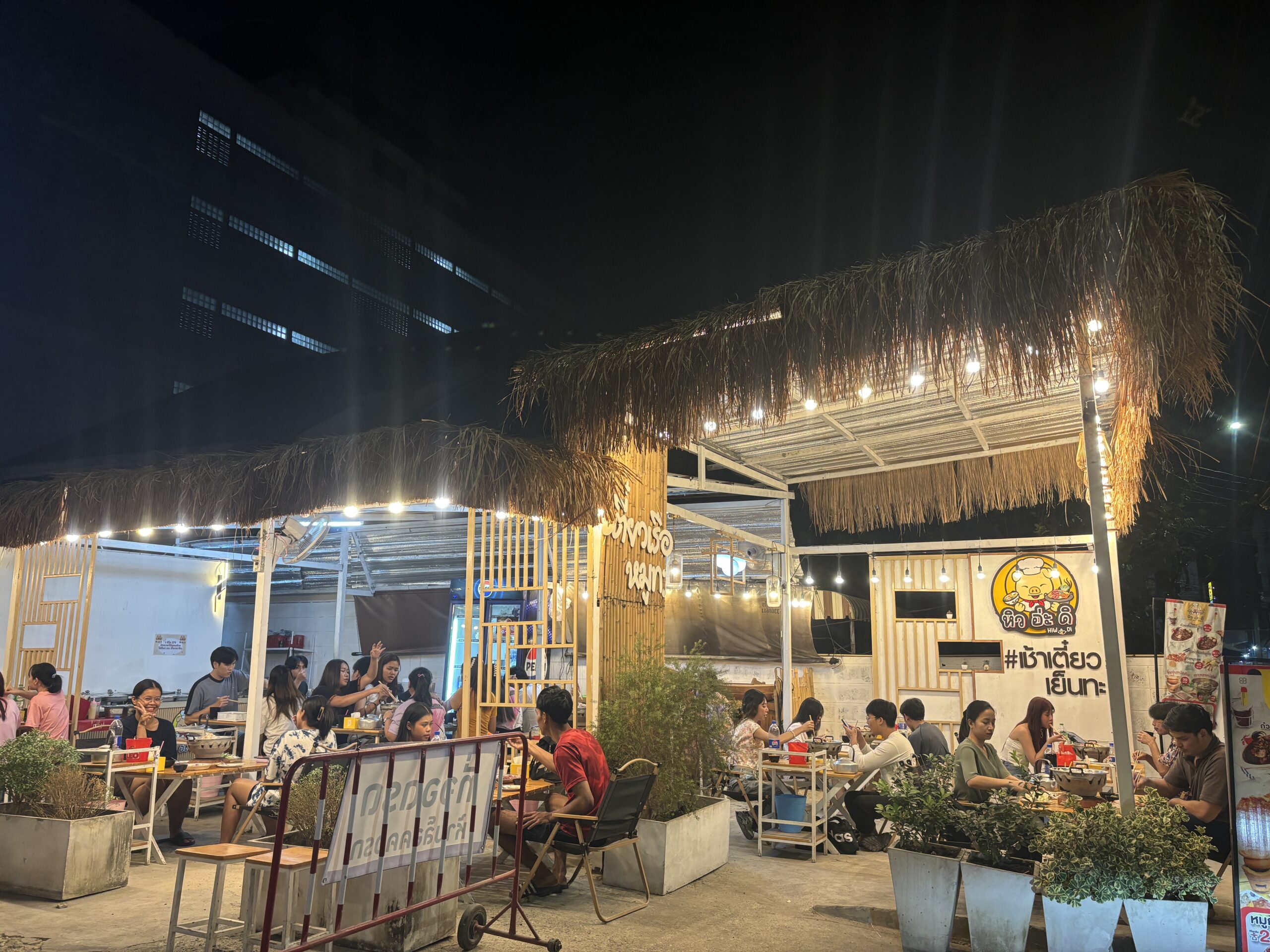 ร้านอาหารกลางแจ้งยามค่ำคืนสไตล์หลังคาหญ้าคา มีลูกค้านั่งทานอาหารหลายโต๊ะ ตกแต่งด้วยไฟสายและตู้คอนเทนเนอร์ ป้ายร้านหิว อิ่ะ ดิ พร้อมแฮชแท็ก #เช้าเตียว เย็นทะ
