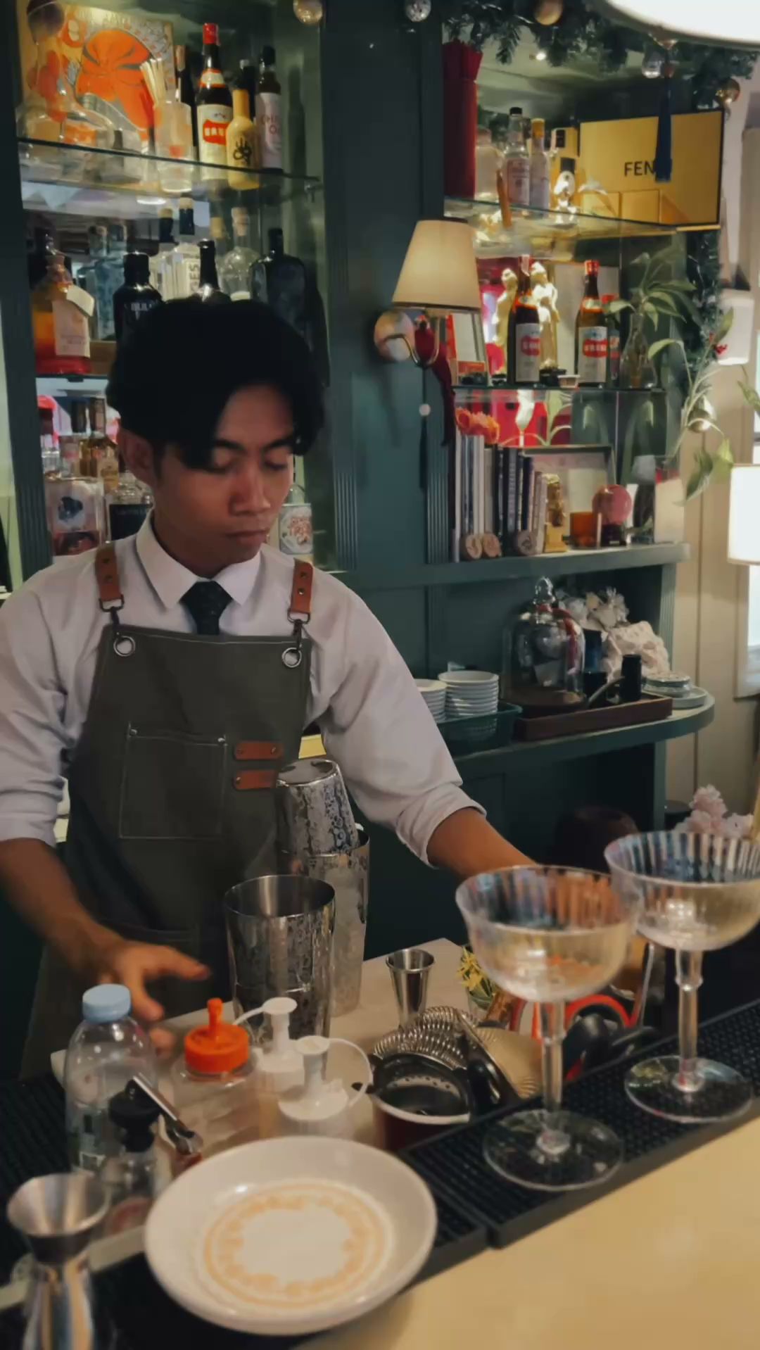 บาร์เทนเดอร์สวมผ้ากันเปื้อนกำลังเตรียมค็อกเทลที่บาร์พร้อมชั้นวางเครื่องดื่มแอลกอฮอล์ด้านหลัง