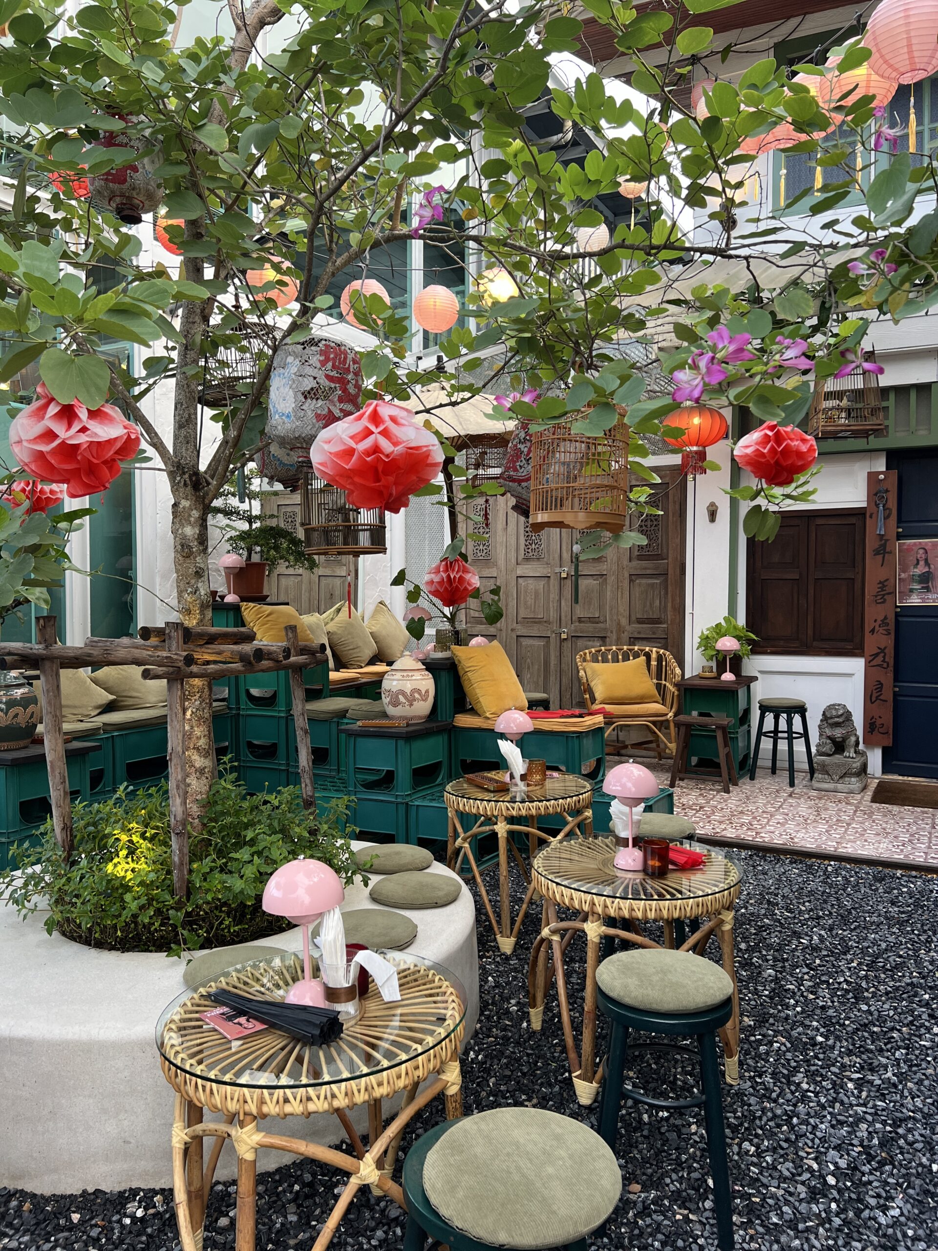 คาเฟ่สไตล์จีนกลางแจ้งตกแต่งด้วยโคมไฟสีแดงและชมพู โต๊ะหวายกระจก เก้าอี้หมอนอิงสีเหลือง ม้านั่งลังเขียว ต้นไม้ใหญ่ และกรงนกไม้แขวนประดับ