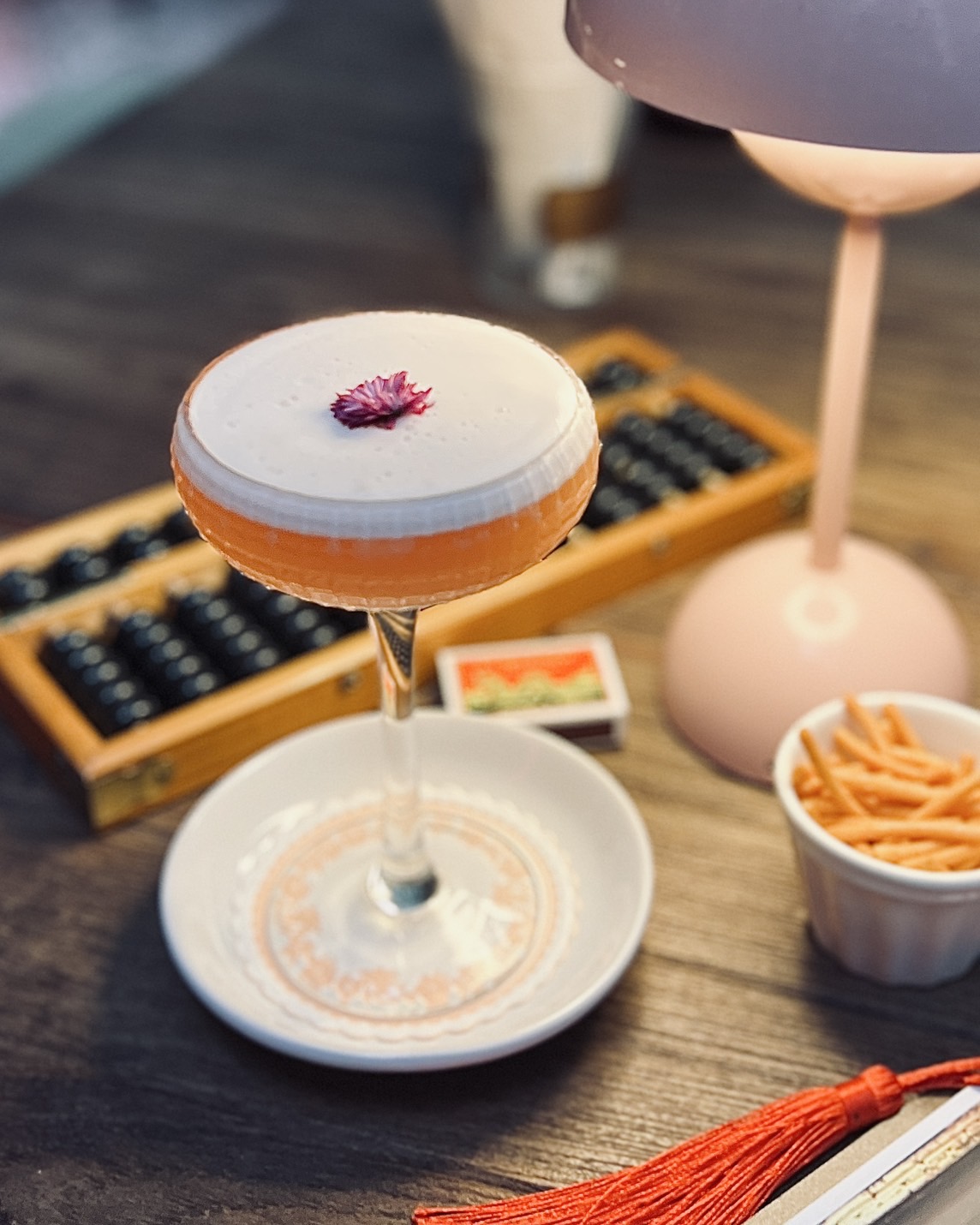 ค็อกเทลสีส้มในแก้วคูเป้ตกแต่งด้วยฟองนุ่มและดอกไม้สีม่วง วางบนจานรองบนโต๊ะไม้พร้อมลูกคิดจีนและโคมไฟสีชมพู