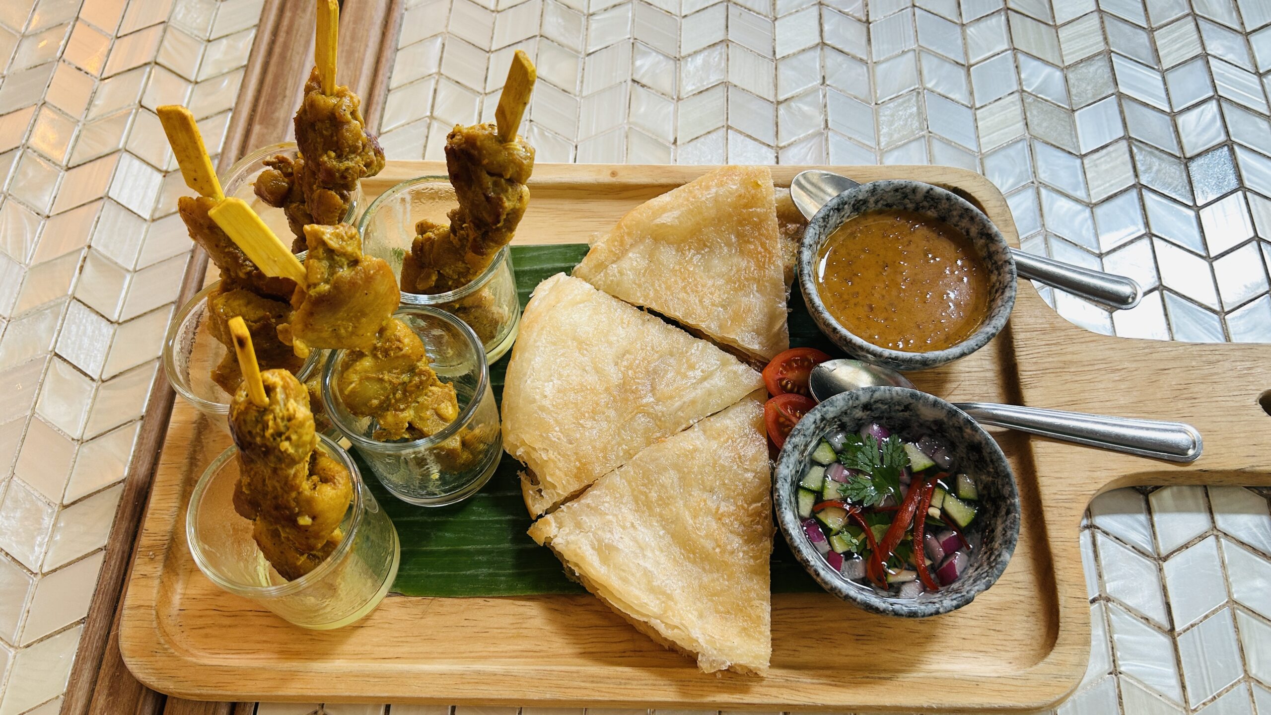 จานไม้เสิร์ฟสะเต๊ะไก่เสียบไม้ในแก้วพร้อมโรตีทอดกรอบ น้ำจิ้มสะเต๊ะ และอาจาดบนใบตอง