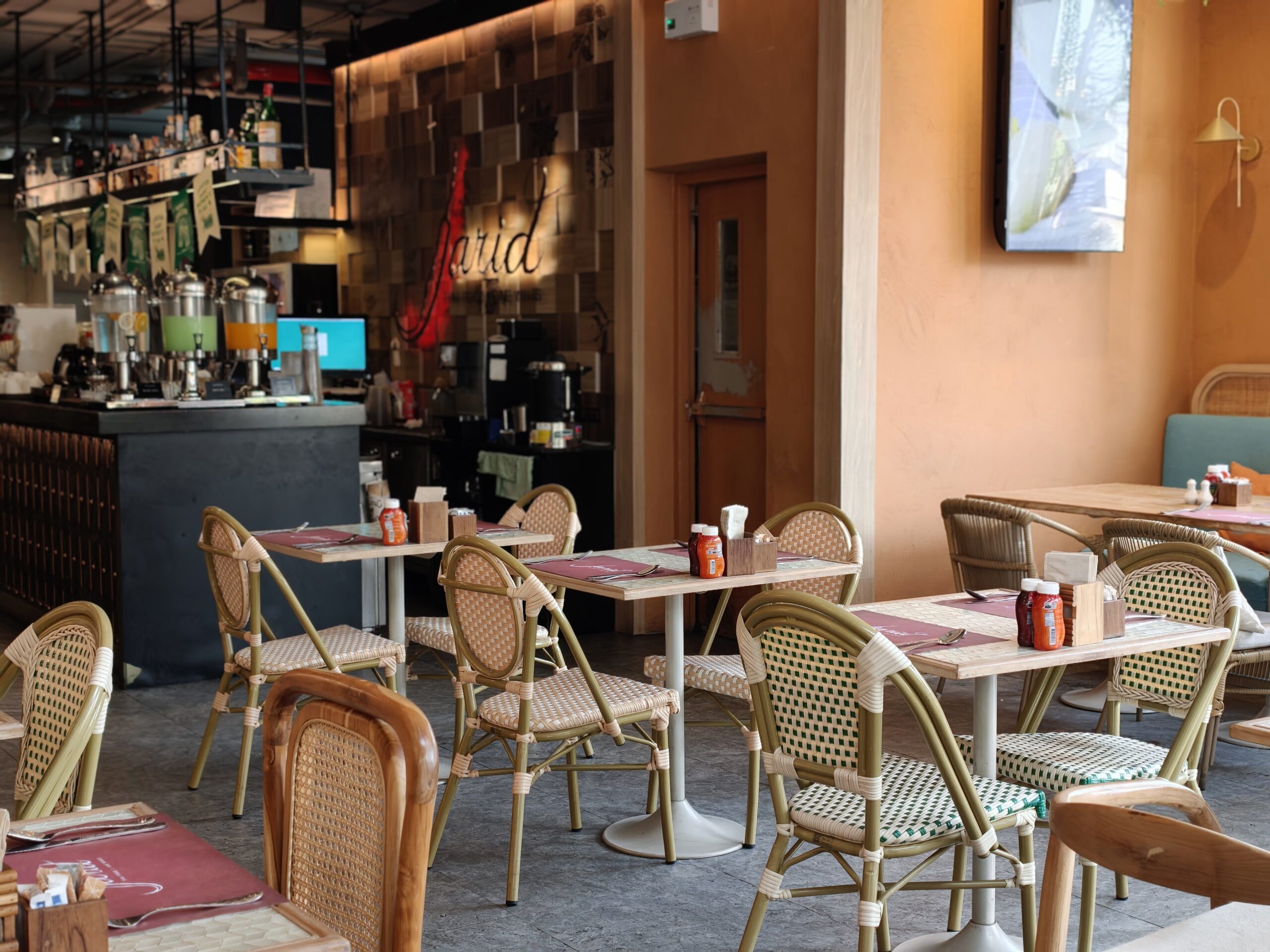 บรรยากาศภายในร้านอาหาร Farid ที่มีโต๊ะและเก้าอี้หวายสไตล์บิสโทร พร้อมเคาน์เตอร์บาร์และเครื่องดื่ม ผนังสีส้มอบอุ่น