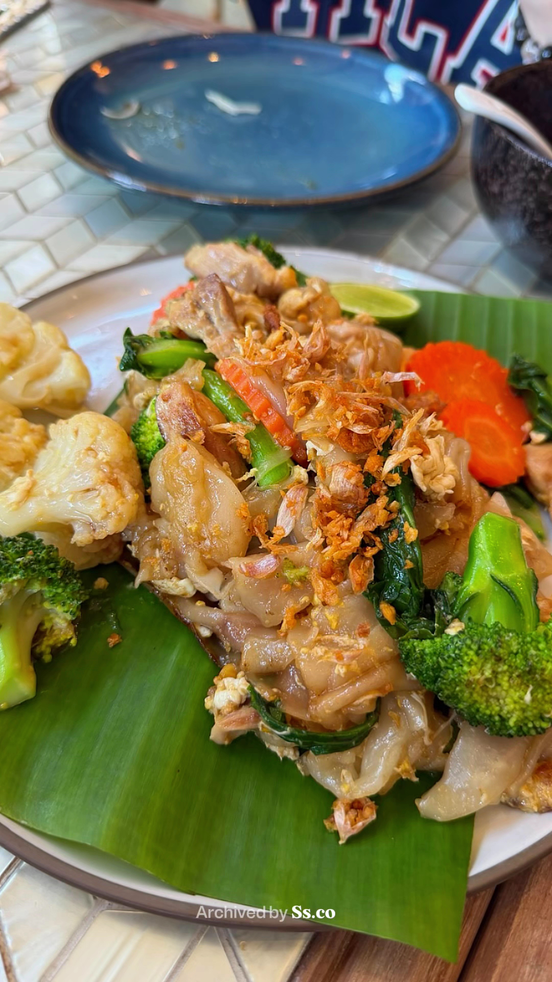 ผัดซีอิ๊วเส้นใหญ่กับไก่และผักบนใบตอง โรยกระเทียมเจียว เสิร์ฟพร้อมบร็อคโคลี่ กะหล่ำดอก และแครอท