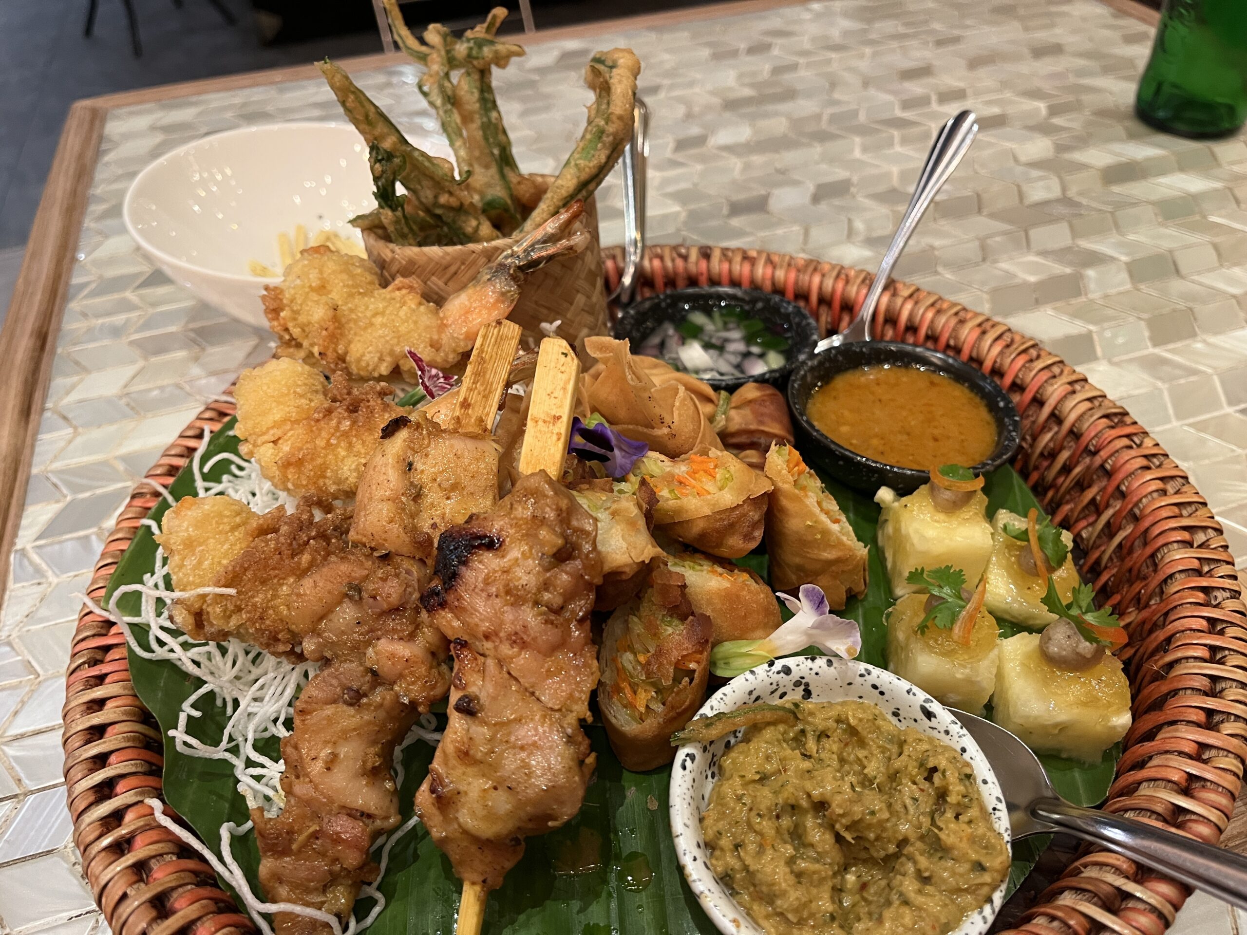 จานเรียกน้ำย่อยรวมมิตรอาหารไทยบนตะกร้าหวาย มีไก่สะเต๊ะ ปอเปี๊ยะทอด ผักทอดกรอบ และน้ำจิ้มหลากชนิด