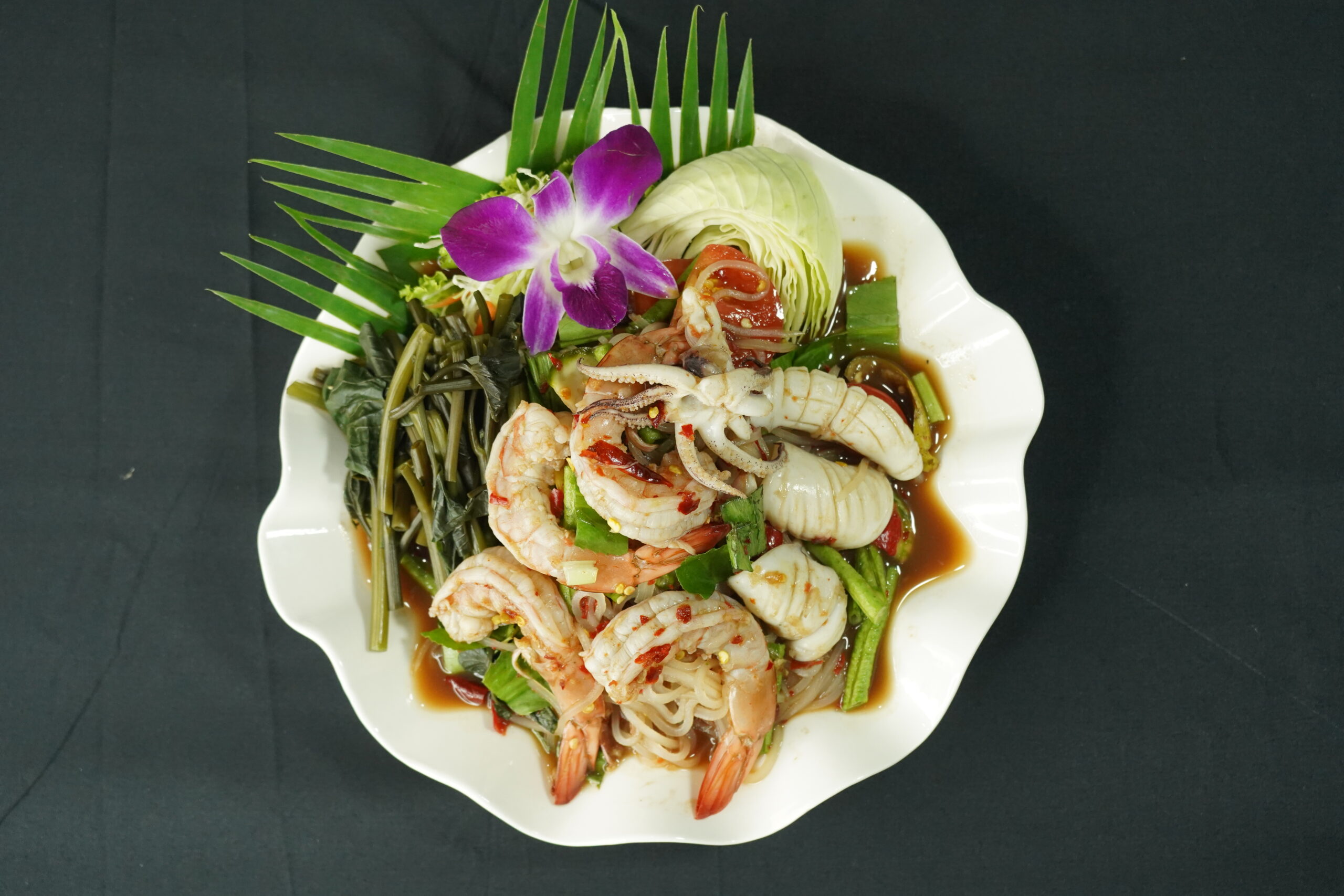 ยำทะเลรวมมิตรจัดจานสวยงามพร้อมกุ้ง ปลาหมึก ผักบุ้ง กะหล่ำปลี ตกแต่งด้วยดอกกล้วยไม้สีม่วงและใบปาล์มบนจานสีขาว