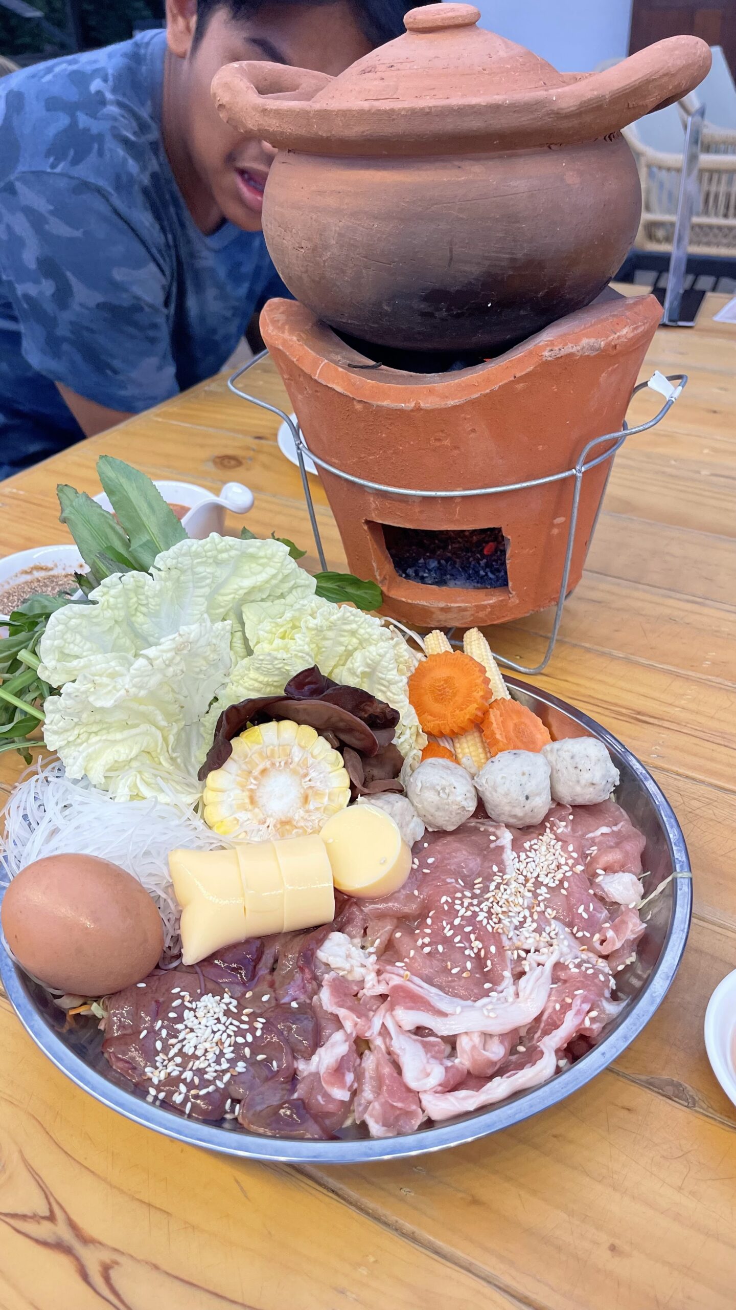 ชุดจิ้มจุ่มหม้อดินพร้อมเนื้อหมูสไลซ์โรยงา ลูกชิ้น ผัก ข้าวโพด วุ้นเส้น เต้าหู้ ไข่ และเตาถ่านหม้อดินบนโต๊ะไม้
