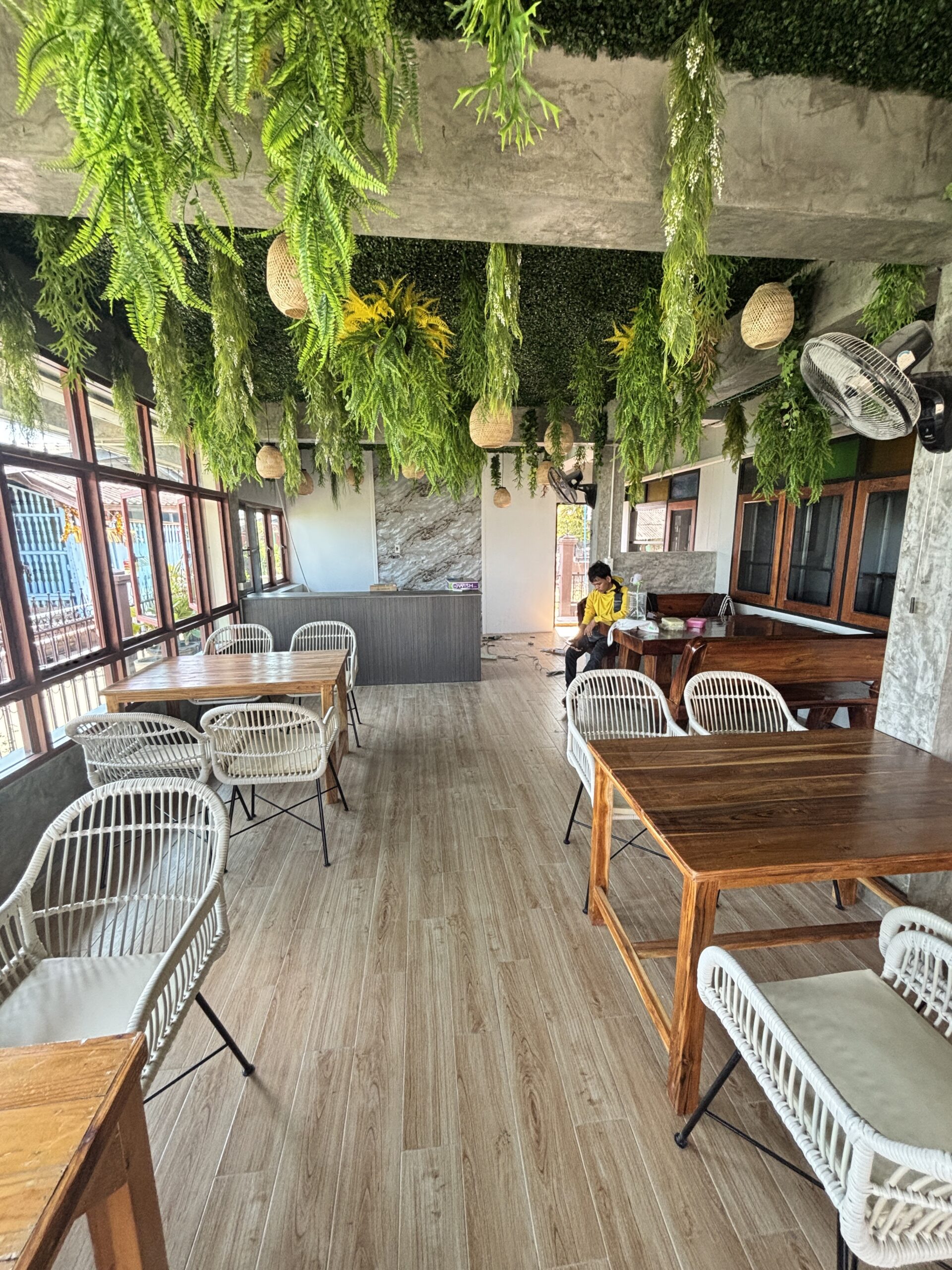 ภายในร้านกาแฟตกแต่งด้วยเฟิร์นเขียวห้อยจากเพดาน โต๊ะไม้และเก้าอี้หวายสีขาว พื้นไม้ลามิเนต บรรยากาศสบายร่มรื่น