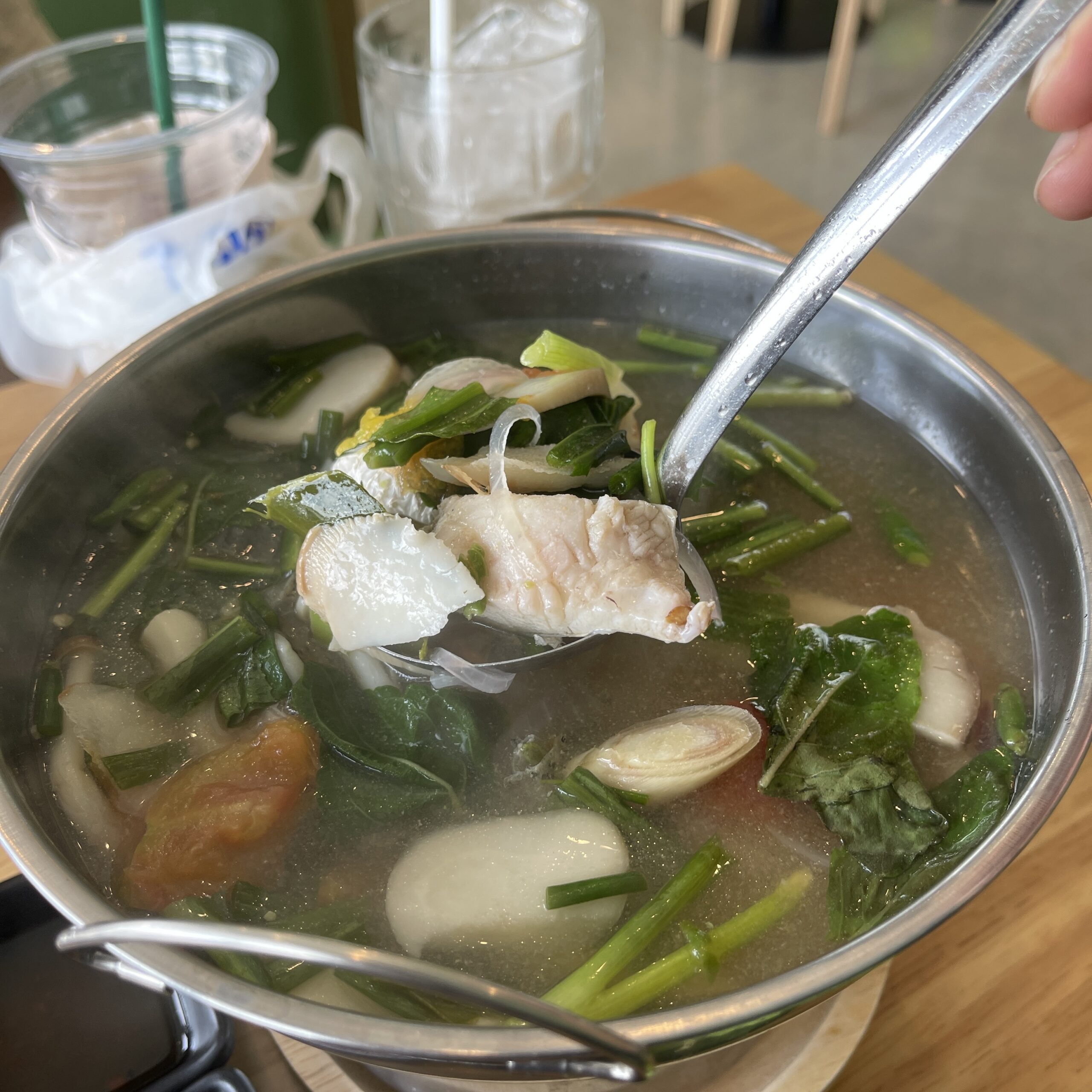 หม้อต้มปลาน้ำใสกับผักบุ้ง มะเขือเทศ และผักชีในหม้อสแตนเลส เสิร์ฟร้อนพร้อมทัพพีตักชิ้นปลาขาวนุ่ม