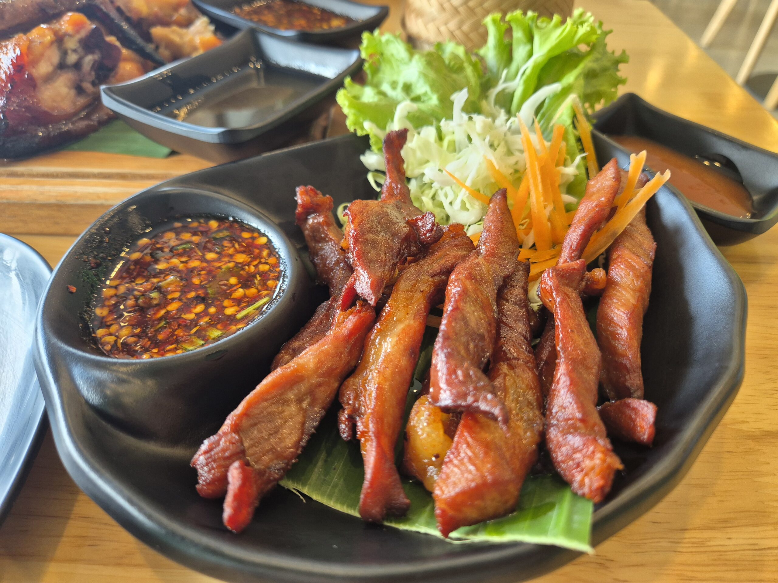 หมูแดดเดียวทอดกรอบเสิร์ฟบนใบตองพร้อมน้ำจิ้มแจ่วและผักสดบนจานสีดำ