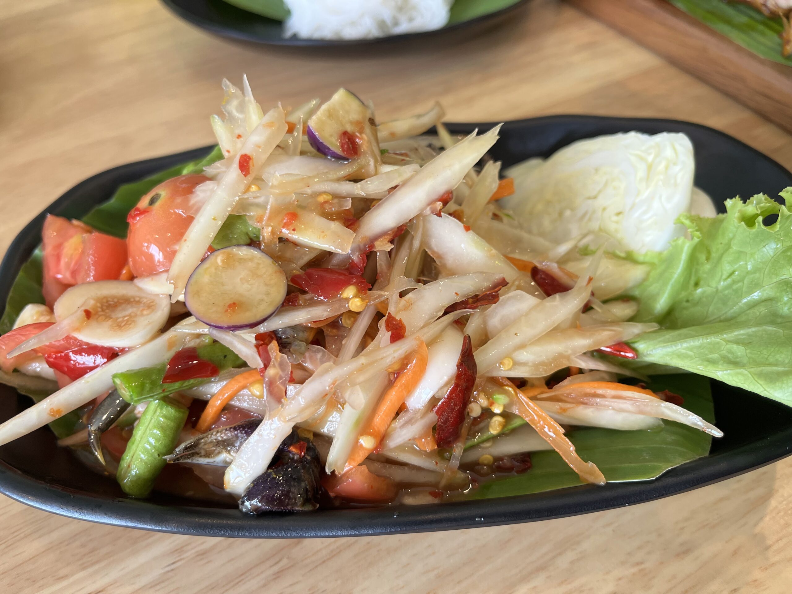 ส้มตำไทยจานใหญ่พร้อมมะเขือเทศ พริกแห้ง ถั่วฝักยาว มะเขือพวง กะหล่ำปลี และผักสด บนจานสีดำ