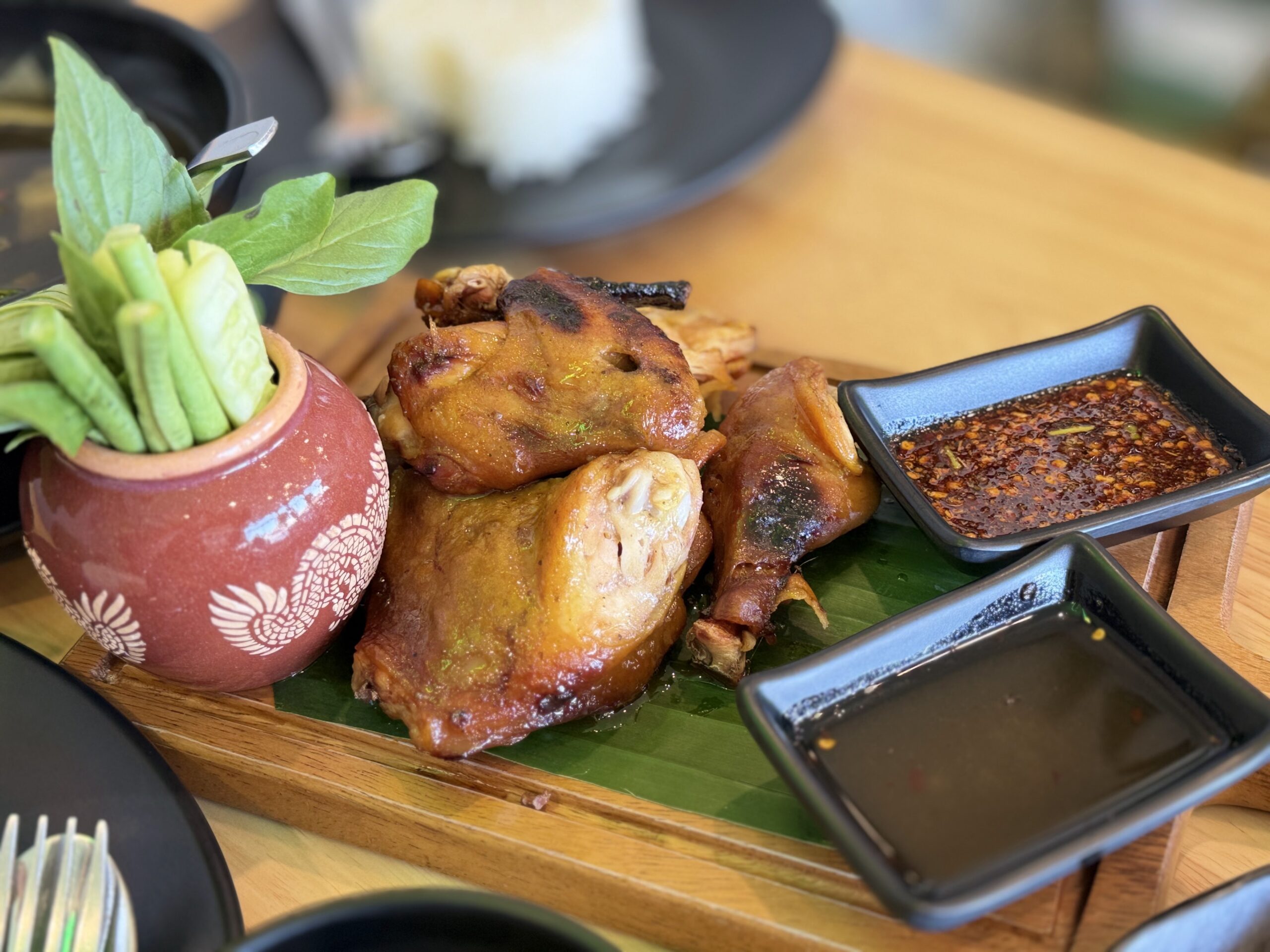 ไก่ย่างเสิร์ฟบนใบตองพร้อมน้ำจิ้มแจ่วและน้ำจิ้มซีอิ๊ว ตกแต่งด้วยผักสดในหม้อดินเผา