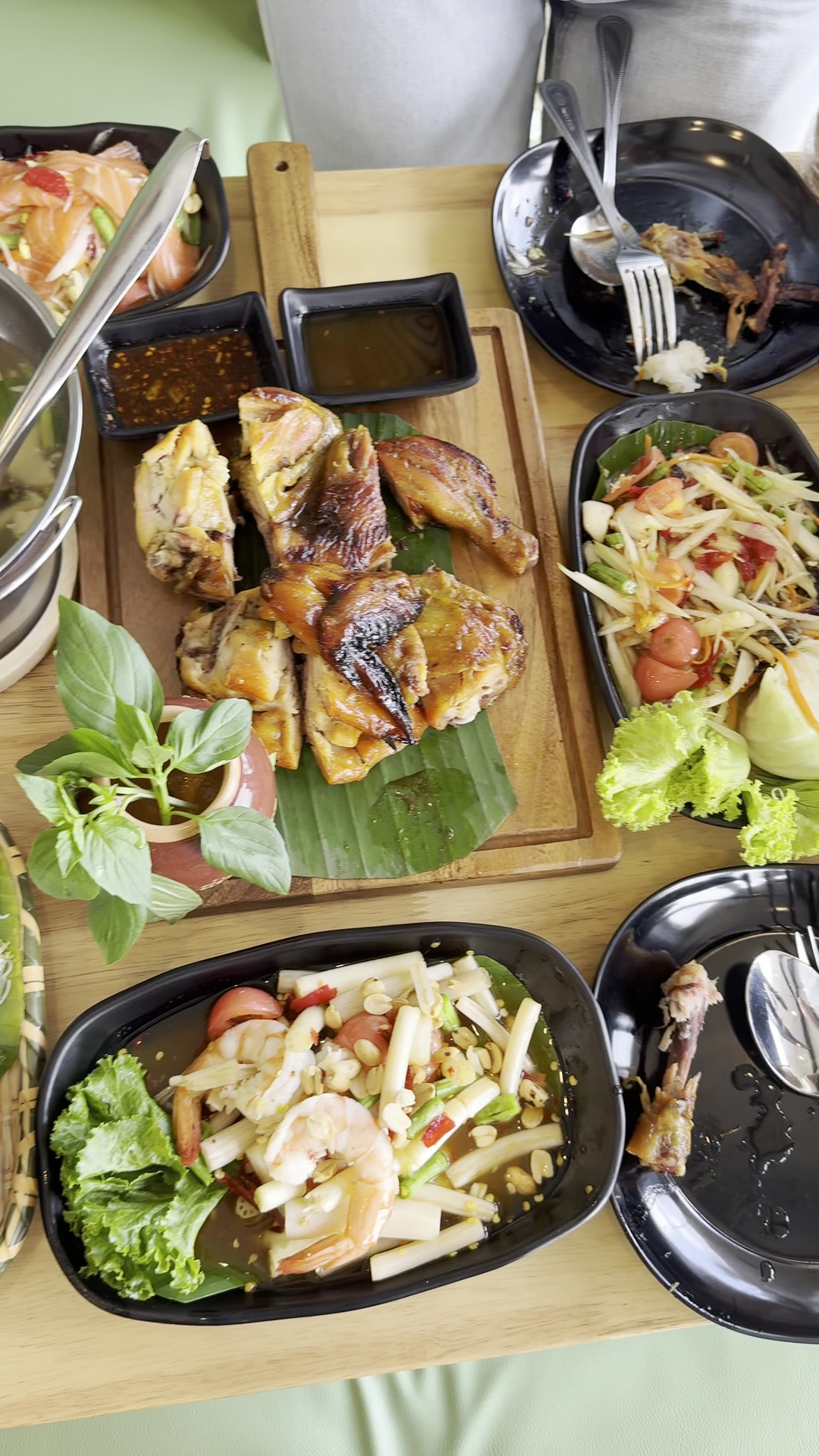 โต๊ะอาหารไทยจัดเต็มมีไก่ย่างบนใบตองพร้อมน้ำจิ้ม ส้มตำ ยำทะเลกับกุ้งสด และสลัดแซลมอน