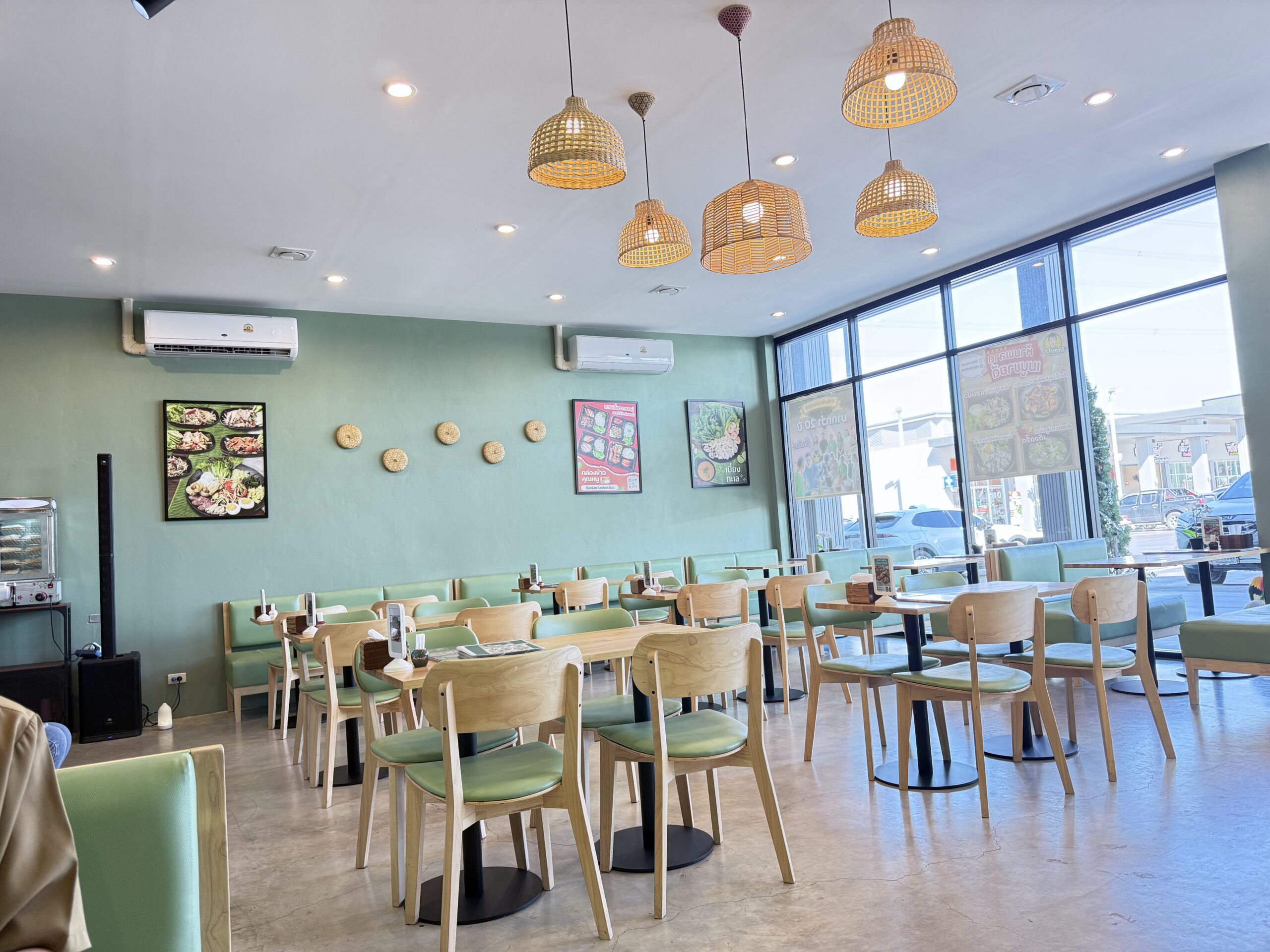 ภายในร้านอาหารไทยตกแต่งโทนสีเขียวมิ้นท์ มีโต๊ะเก้าอี้ไม้เบาะสีเขียว โคมไฟสานไม้ไผ่ห้อยจากเพดาน ผนังประดับรูปเมนูอาหารและงานตกแต่ง พร้อมหน้าต่างกระจกบานใหญ่