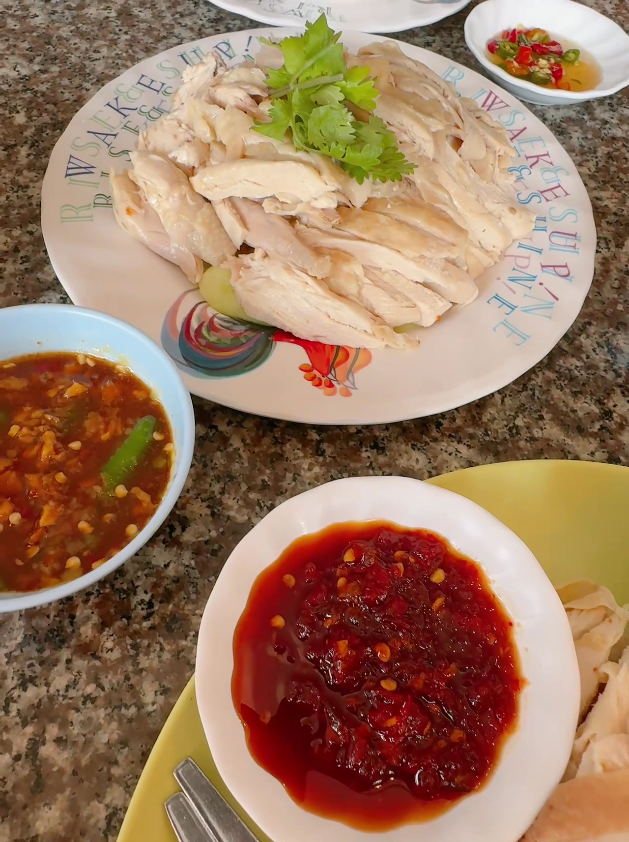 ข้าวมันไก่ต้มหั่นเป็นชิ้นบนจานพร้อมผักชี เสิร์ฟกับน้ำจิ้มพริกแดง น้ำจิ้มถั่วเหลือง และพริกดองน้ำส้ม